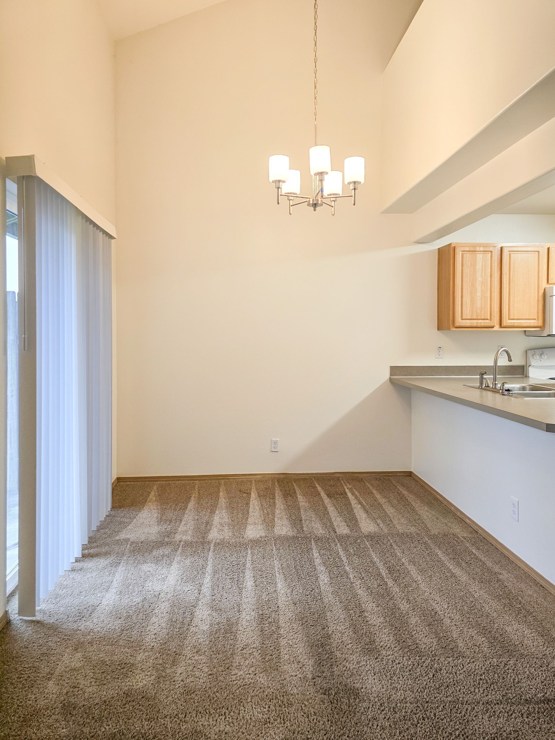 Empty dining area with beige walls, carpet, sliding glass door, and adjacent kitchen counter with cabinets