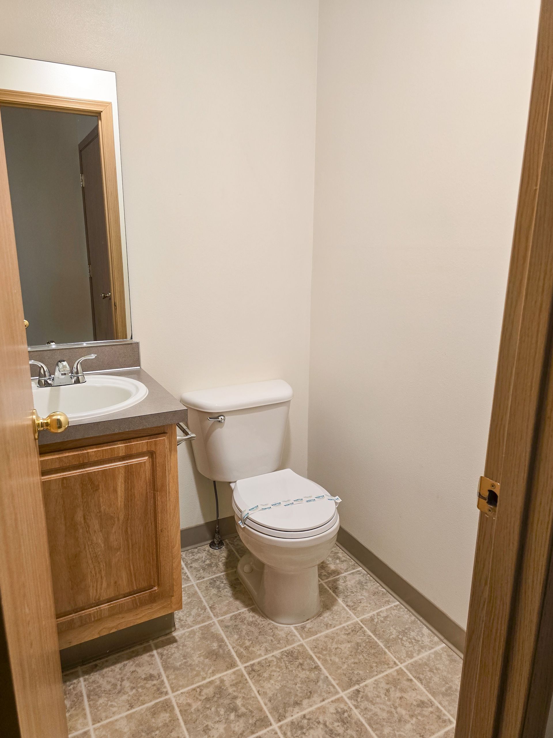 Small bathroom with toilet, sink, mirror, wood vanity, and beige tile floor.