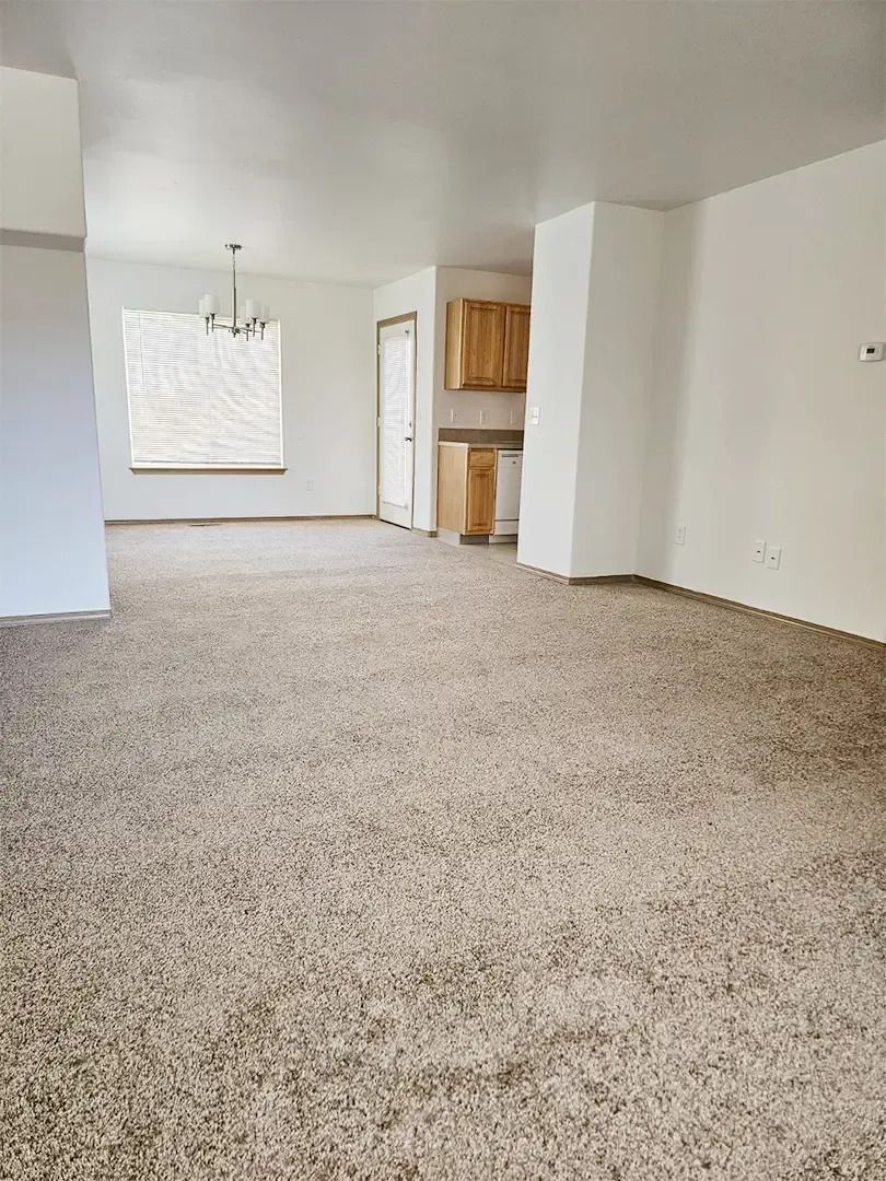 Empty beige-carpeted living room with white walls, a chandelier, and a small kitchen opening.