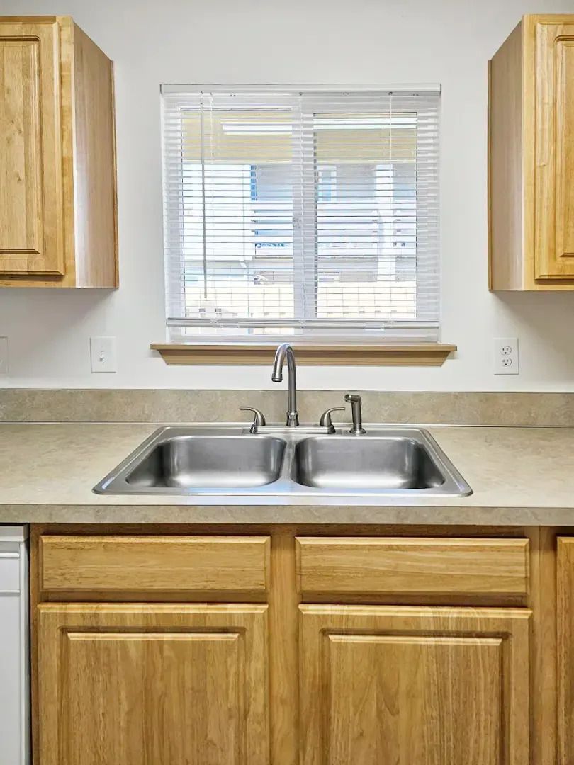 Kitchen sink with double basin, light wood cabinets, and a window with blinds above the counter