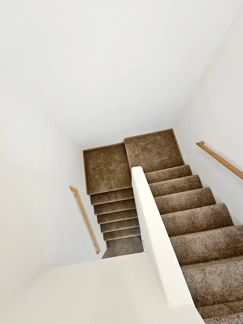 Carpeted staircase with white walls and wooden handrails, viewed from the top landing.