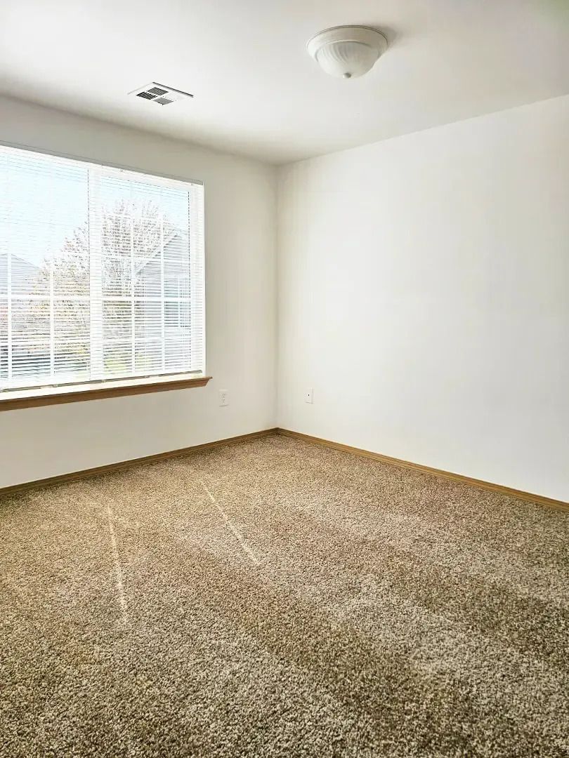 Empty carpeted bedroom with white walls, a window with blinds, and a ceiling light.