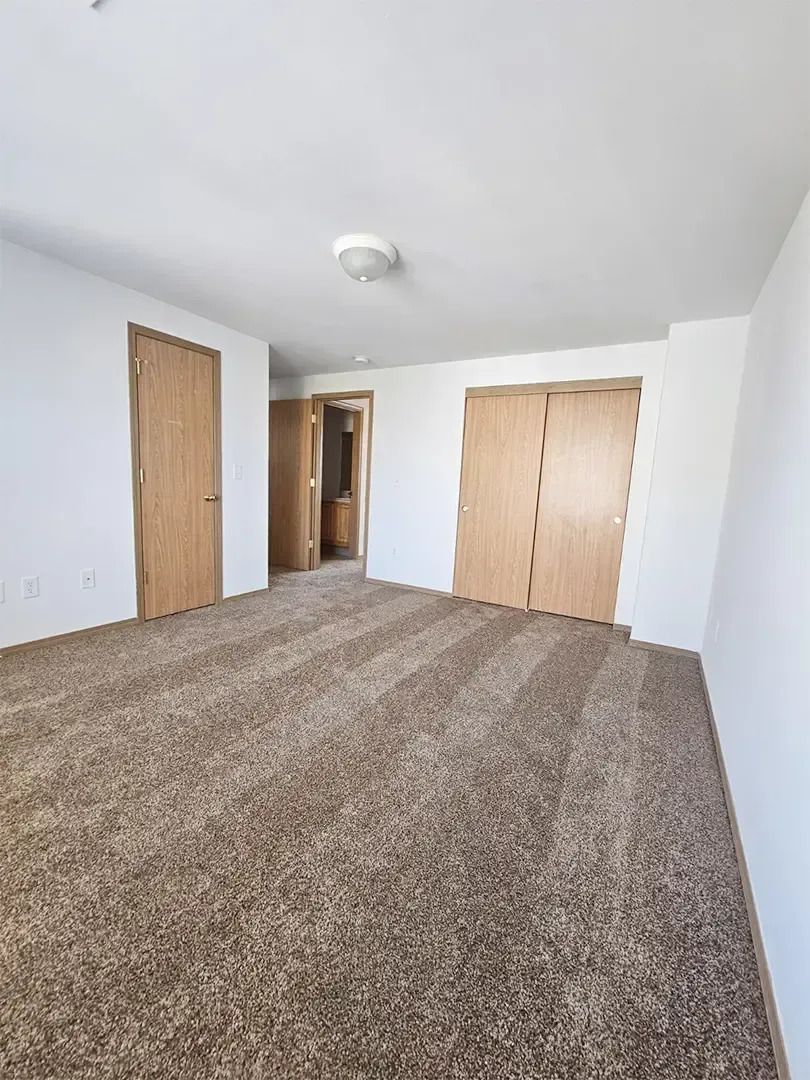 Empty carpeted bedroom with white walls and two wooden doors, viewed from a corner