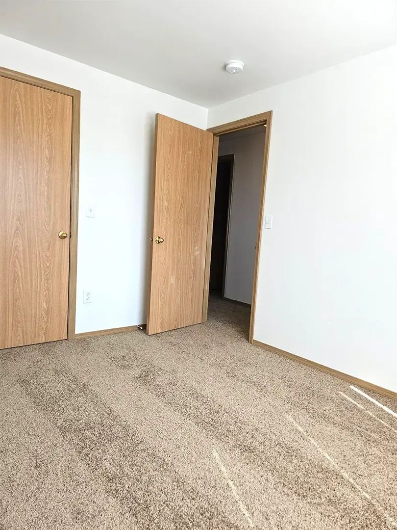 Empty carpeted bedroom with white walls and two wooden doors, one open to a hallway.