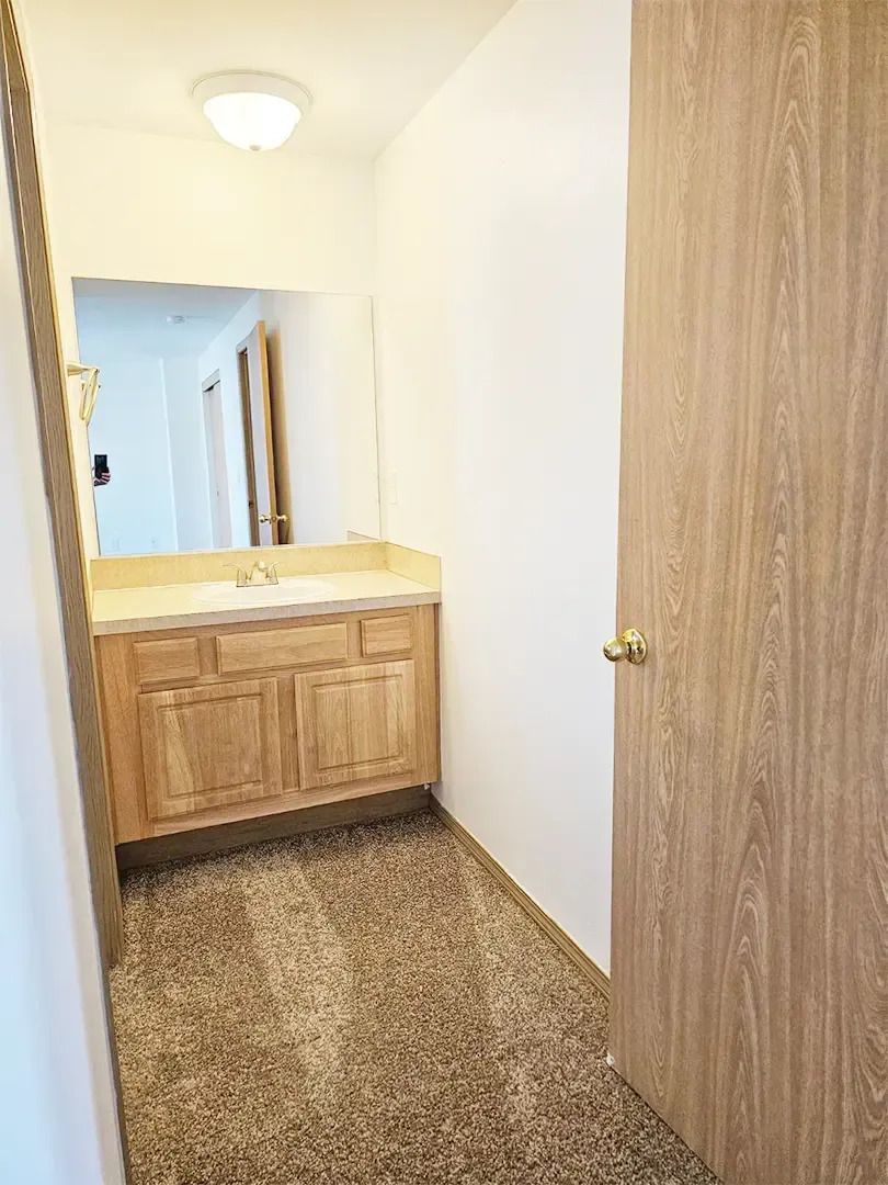 Narrow hallway with wooden vanity, mirror, speckled floor, and closed door beside it.