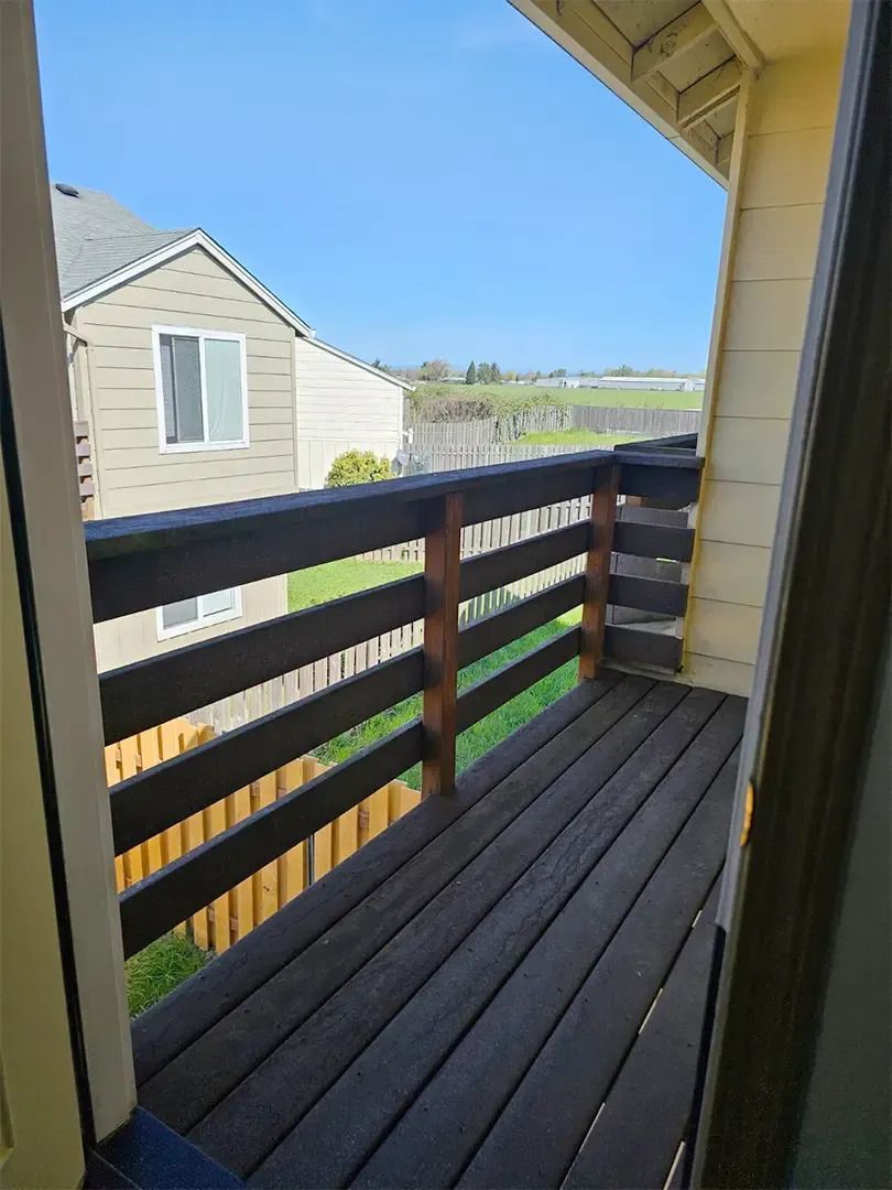Narrow dark wooden balcony with railing, viewed from a doorway, overlooking a grassy yard and neighboring house.