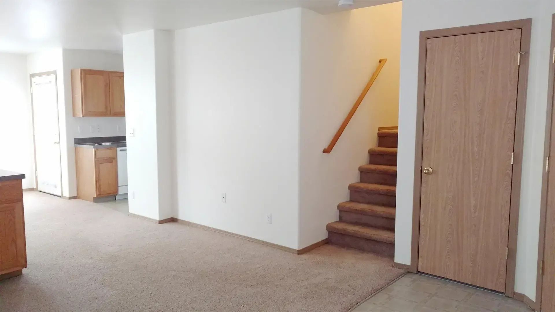 Empty living room with beige carpet, white walls, a kitchen opening, and stairs leading to an upper floor.