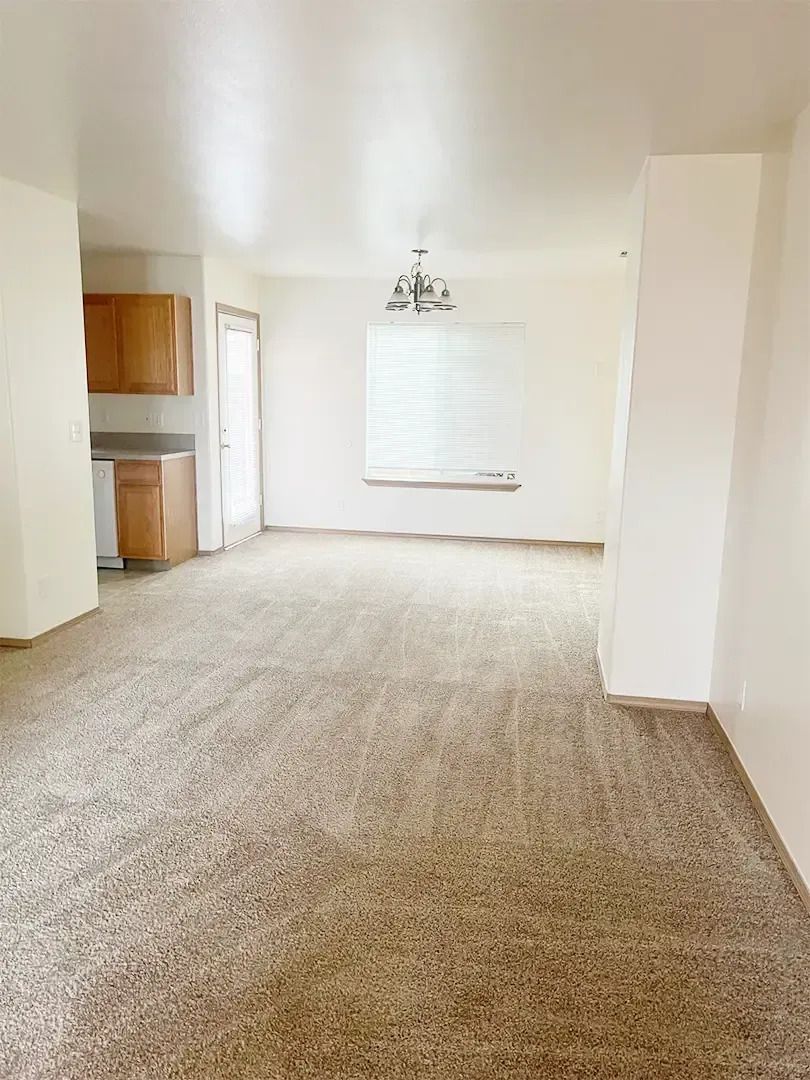 Empty living room with beige carpet, white walls, kitchen nook, and a ceiling light fixture.