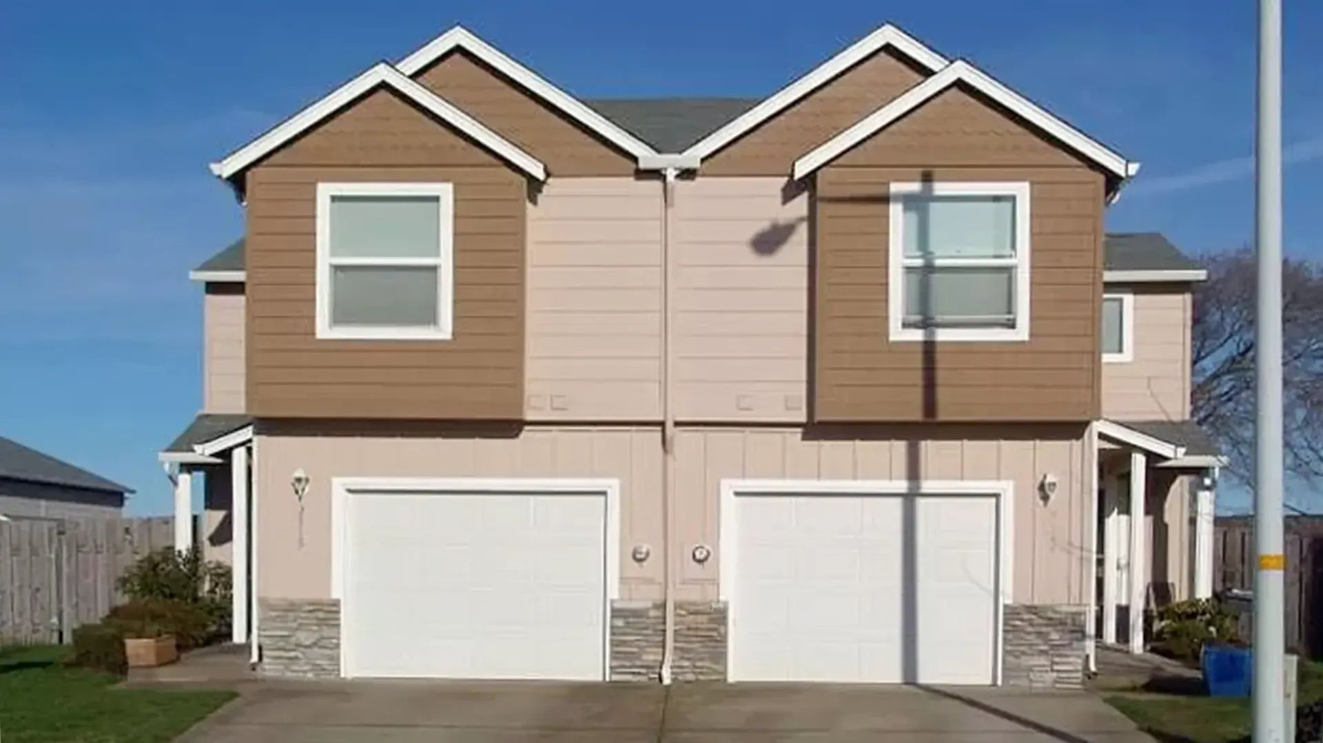 Two-story townhouse with tan siding, white garage doors, and a blue sky background
