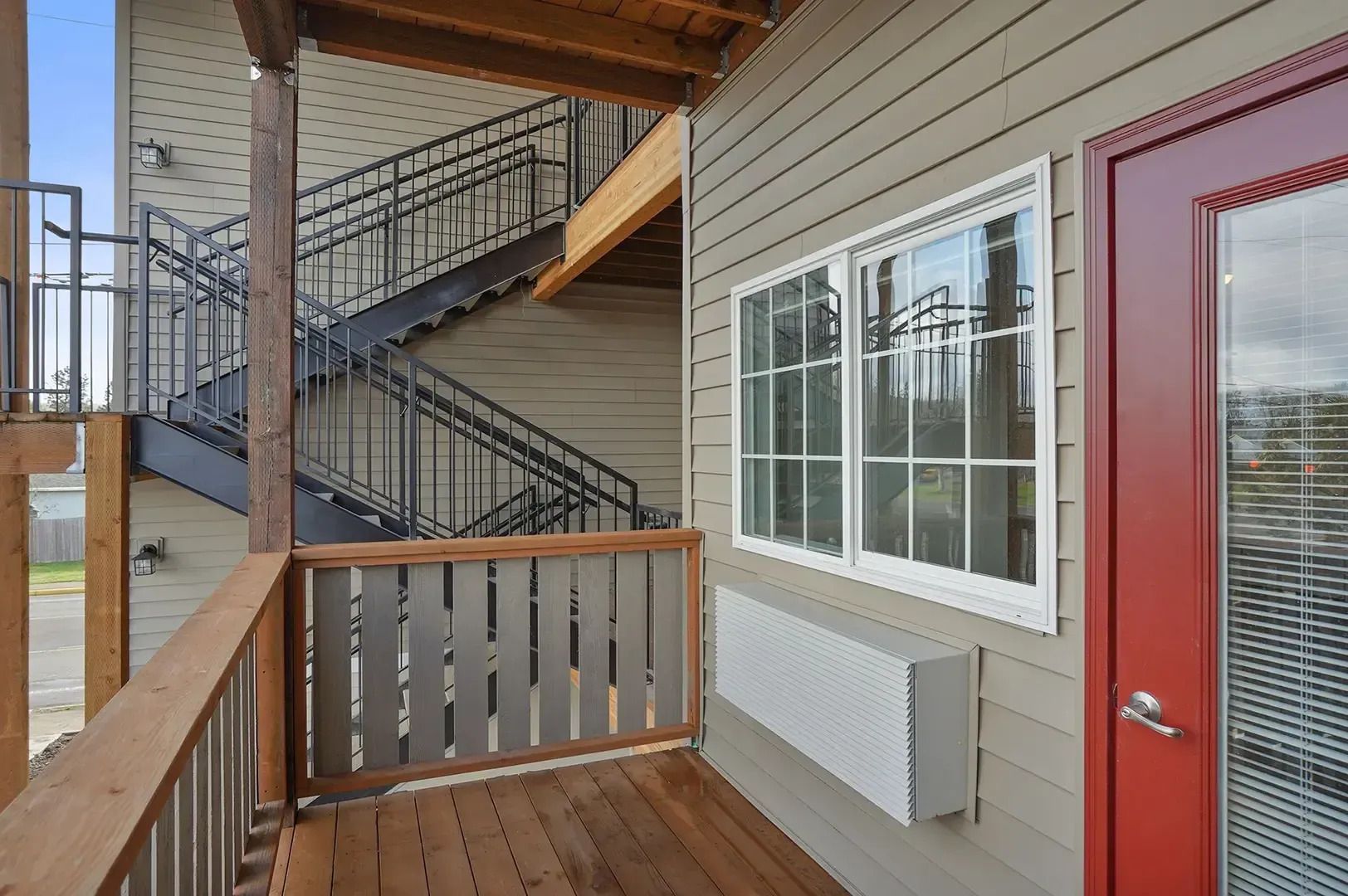 Apartment balcony with wooden floor, beige siding, red door, white window, and metal exterior stairs