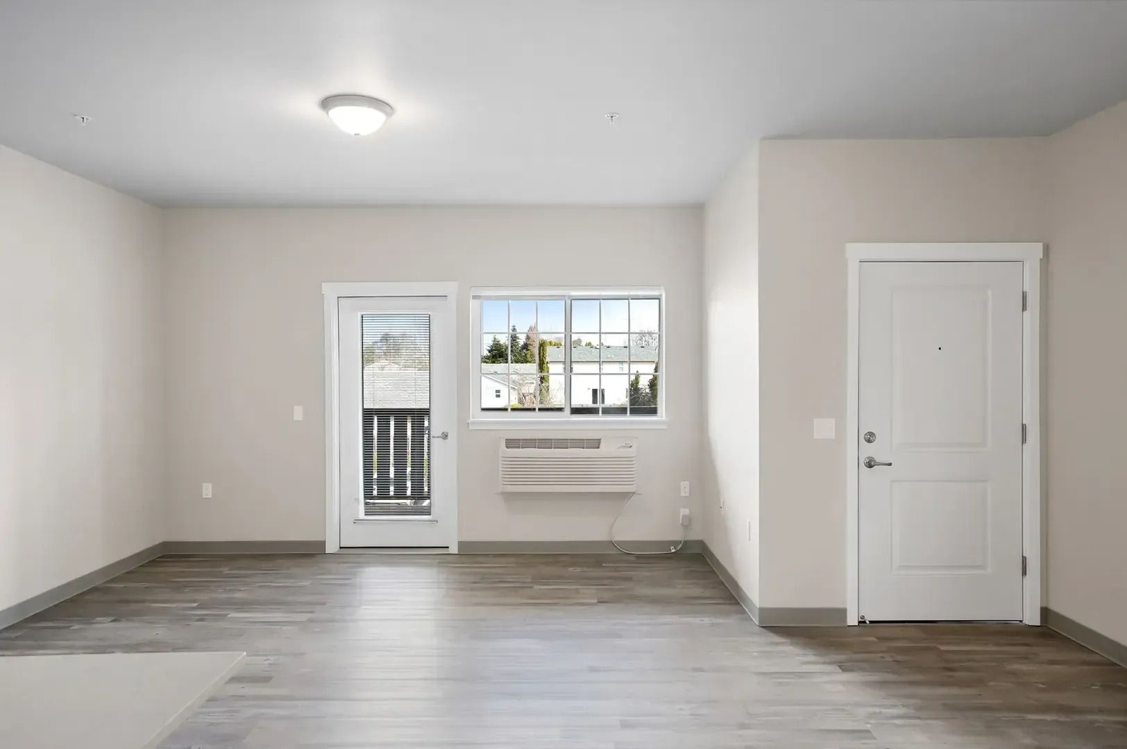 Empty modern apartment room with light gray floors, white walls, a window, and a door to a balcony.