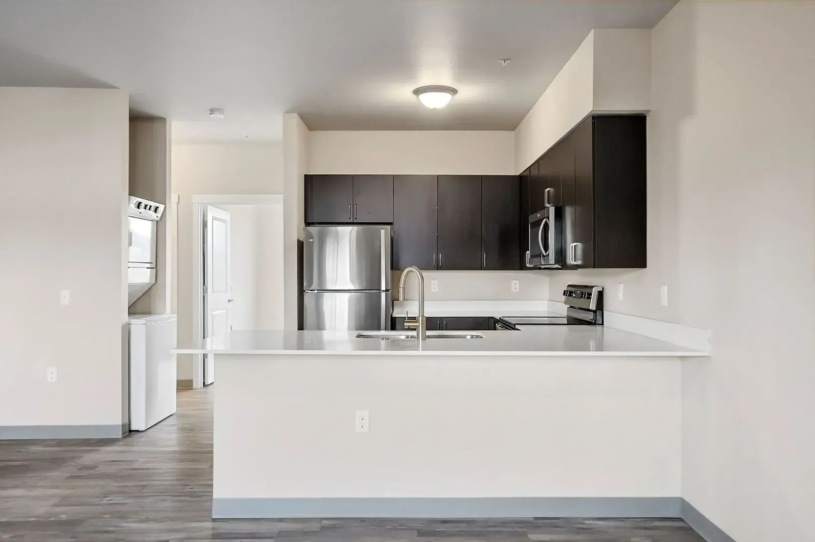 Bright modern kitchen with white counters, dark cabinets, stainless steel appliances, and gray flooring.