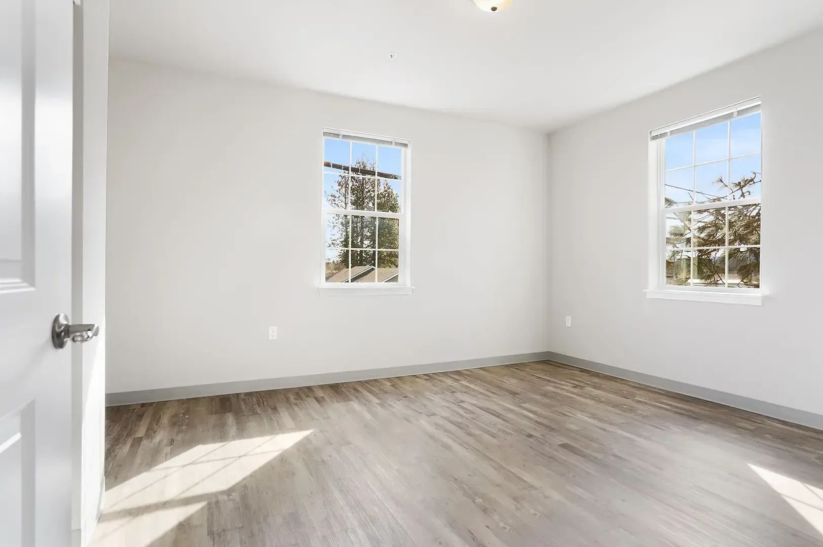 Empty white bedroom with light wood flooring and two windows bringing in sunlight