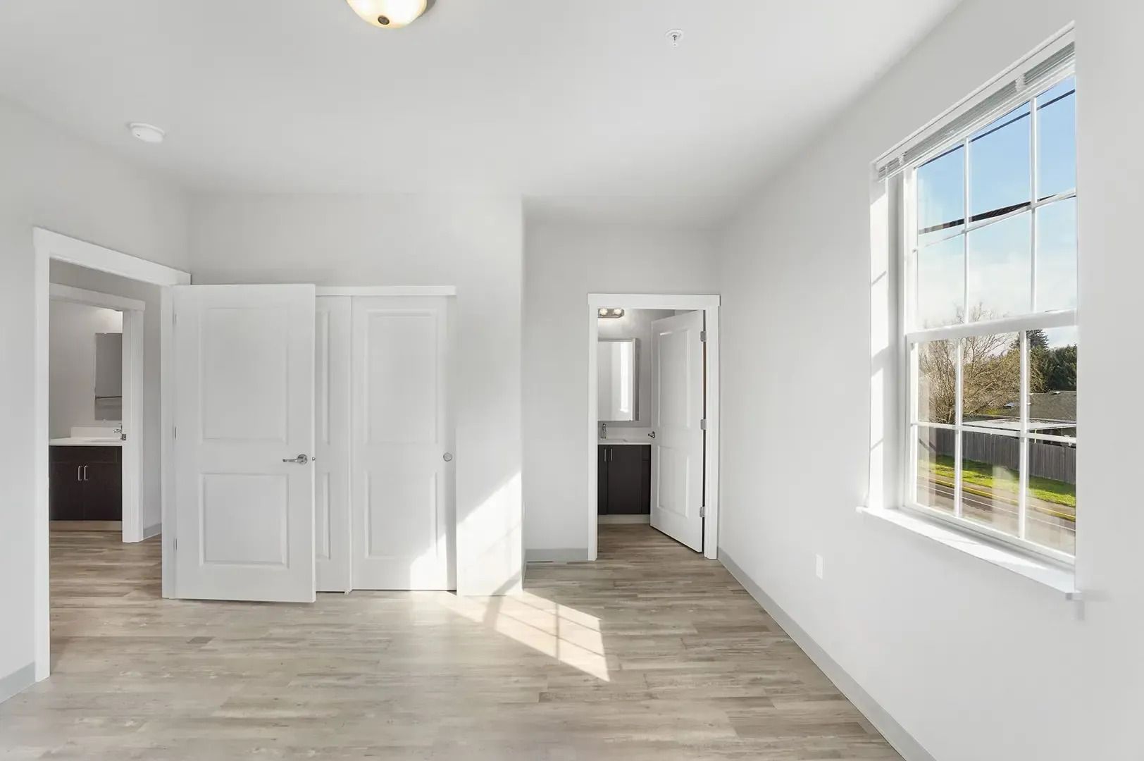 Bright white empty bedroom with two doors and a large window letting in sunlight