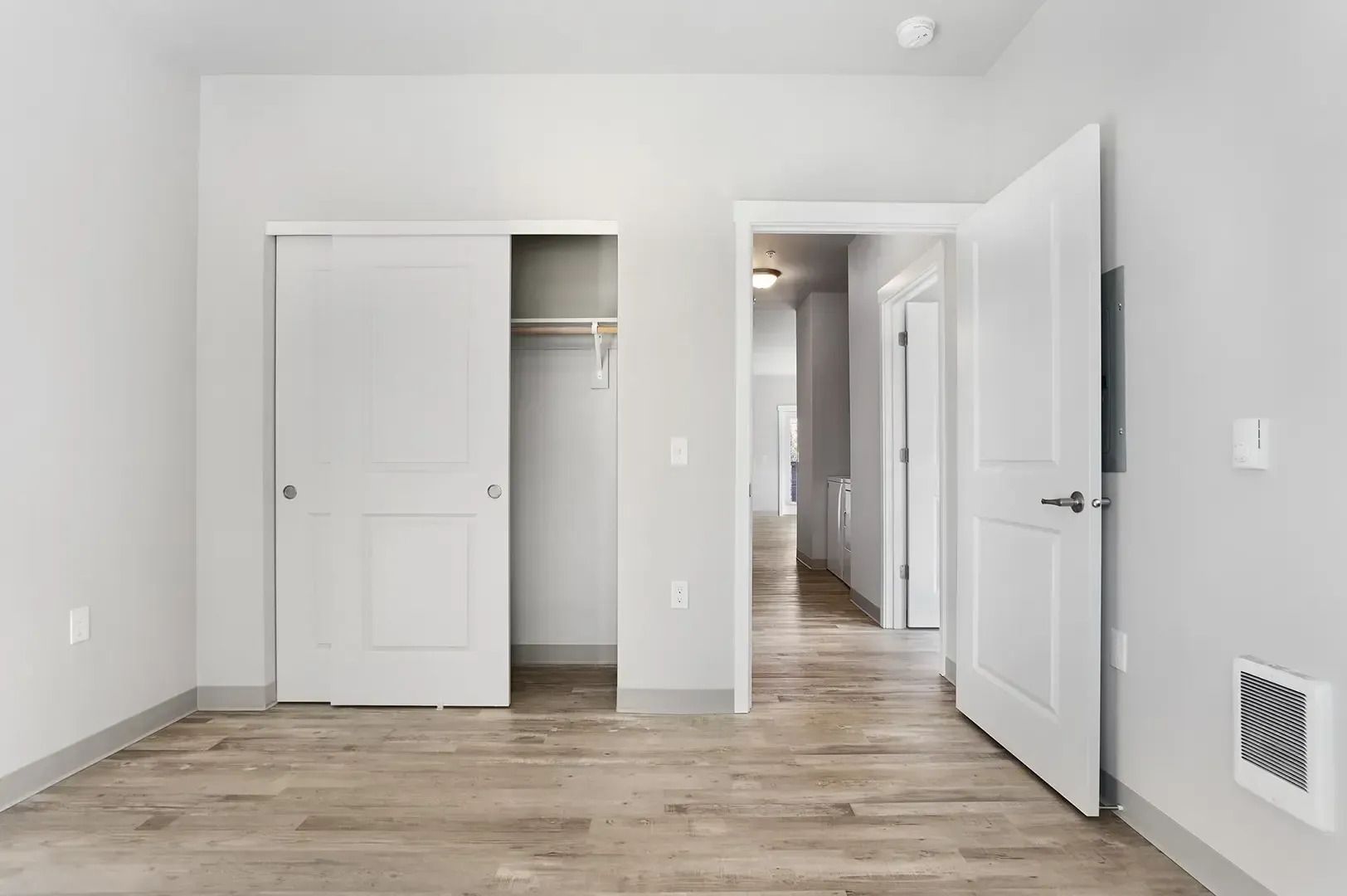 Empty white bedroom with open closet and door, light wood flooring, and a wall vent.