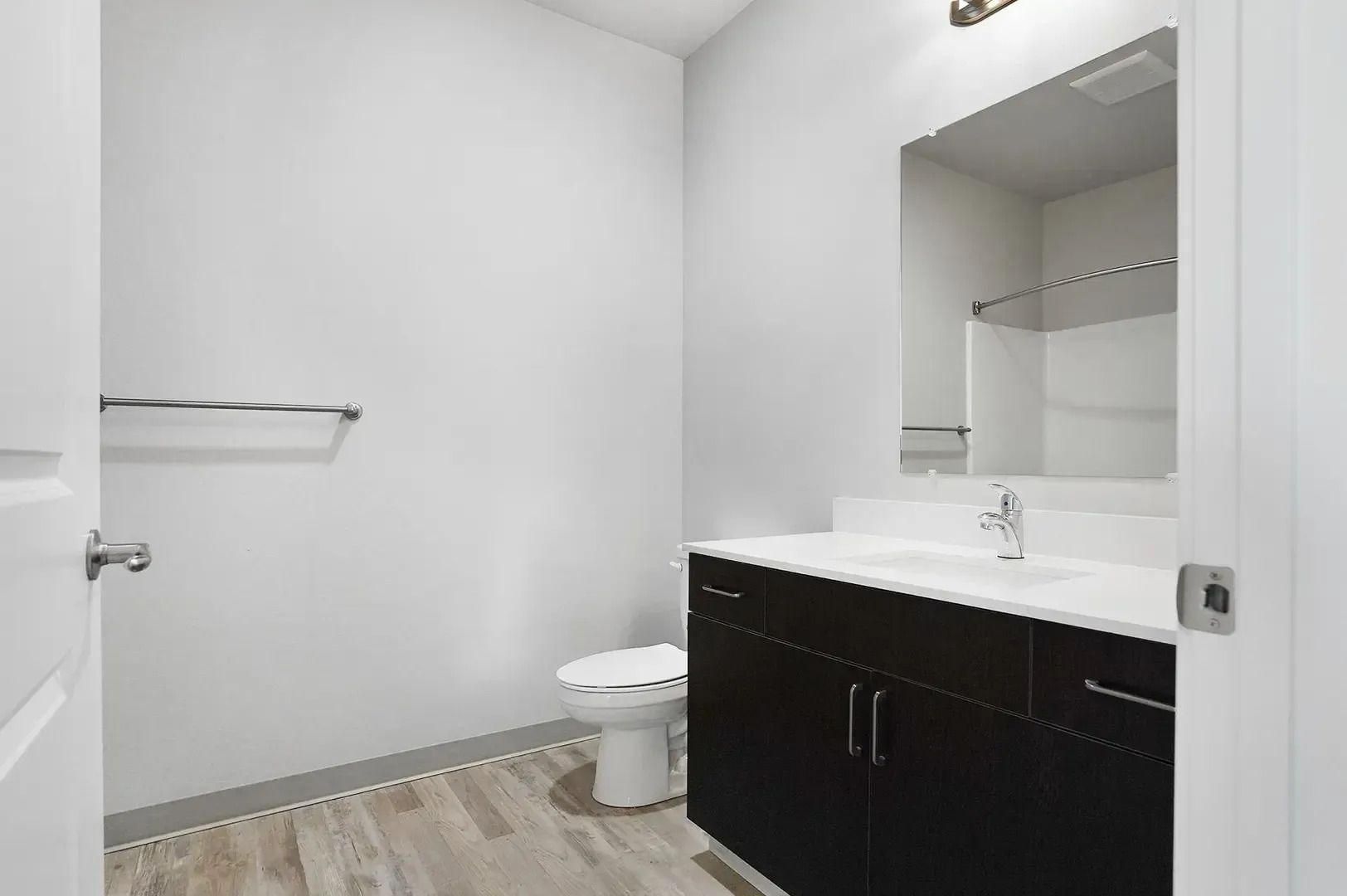 Empty modern bathroom with white walls, dark vanity, mirror, toilet, and light wood flooring