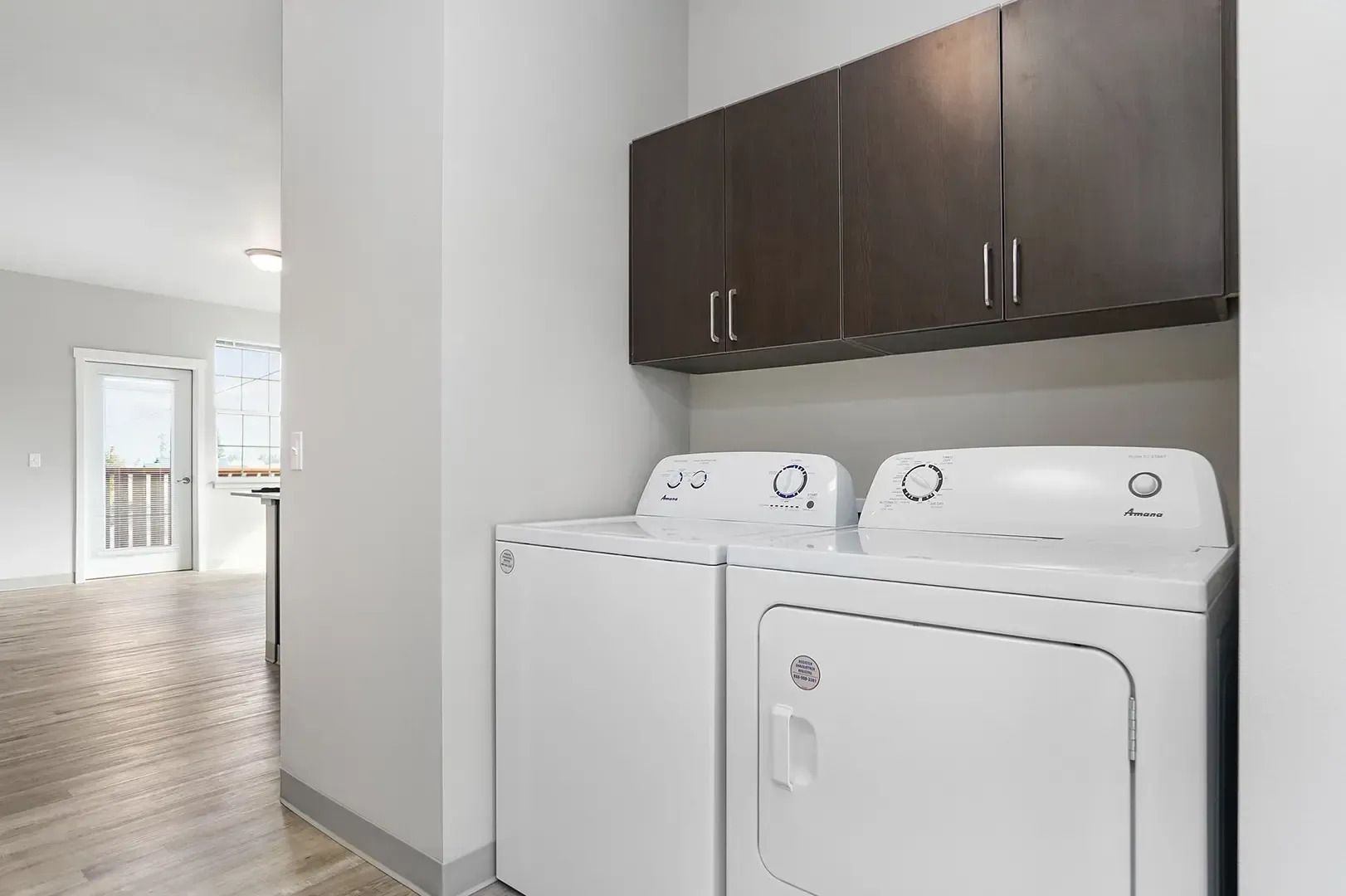 Laundry room with white washer and dryer beneath dark wood cabinets in a bright hallway