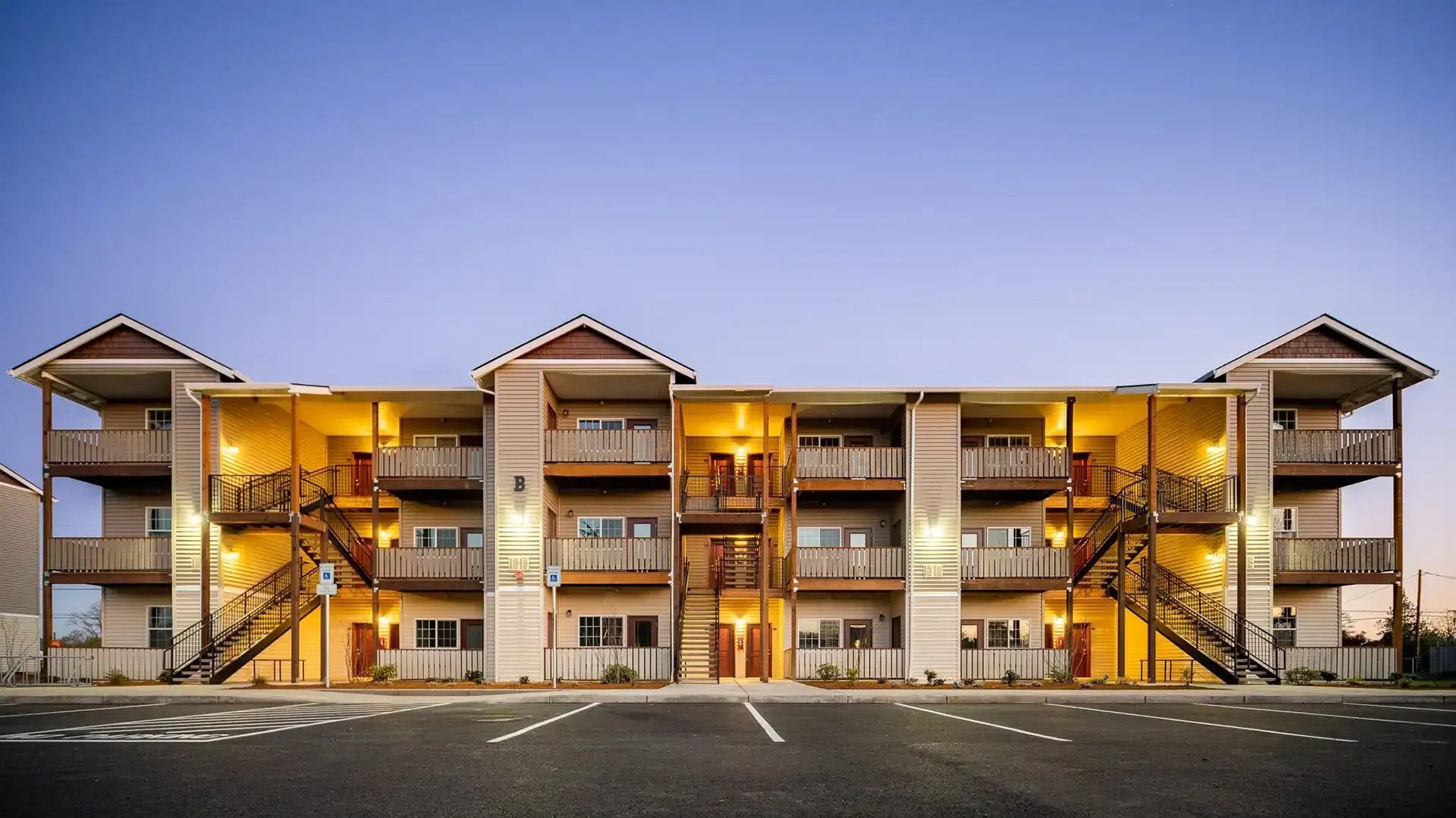 Two-story apartment complex at dusk with lit balconies and staircases, viewed from a parking lot