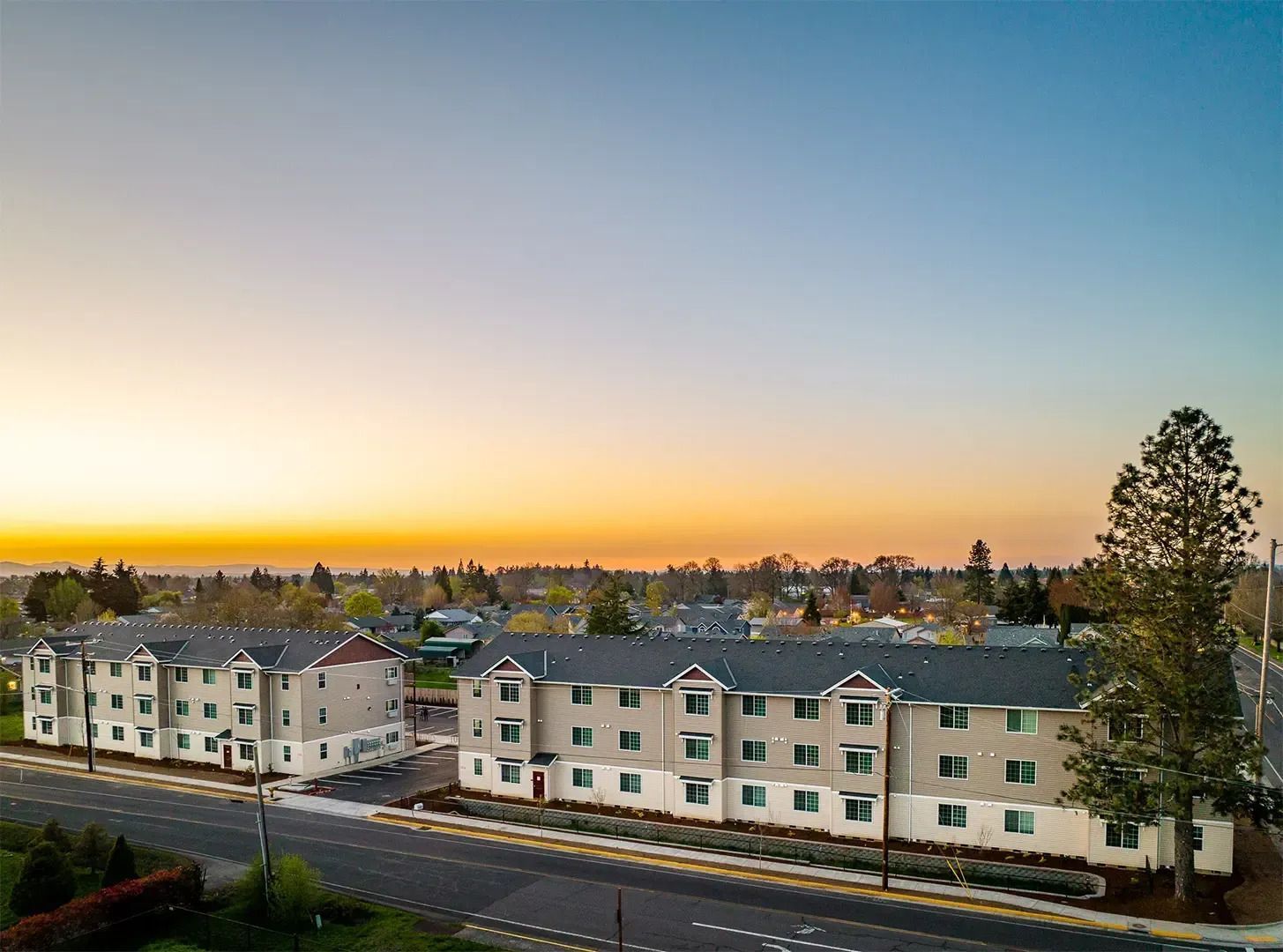 Apartment complex at sunset with a road in front and trees against a clear sky
