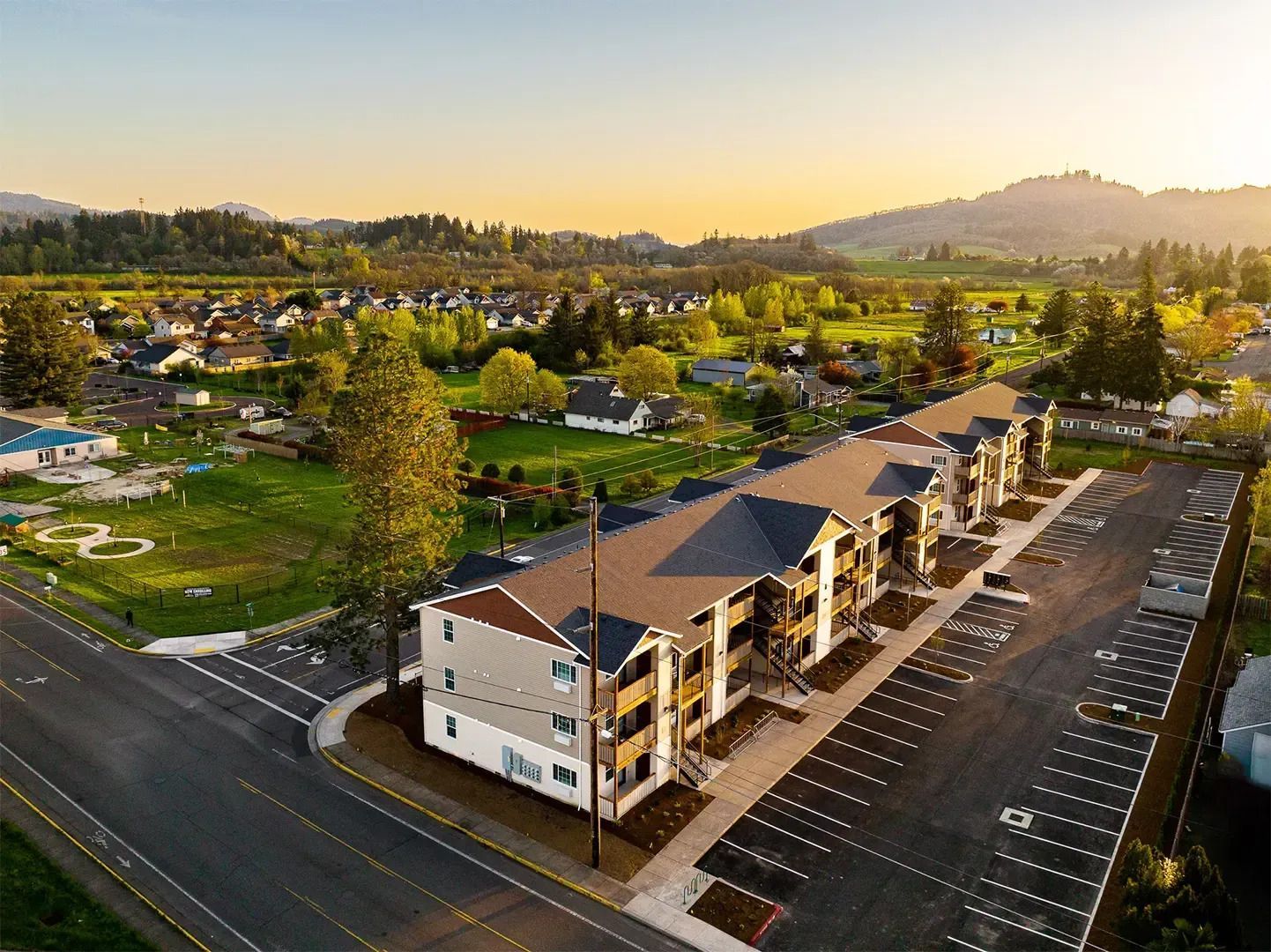 Aerial view of a suburban apartment complex at sunset, with parking lot and green hills beyond.