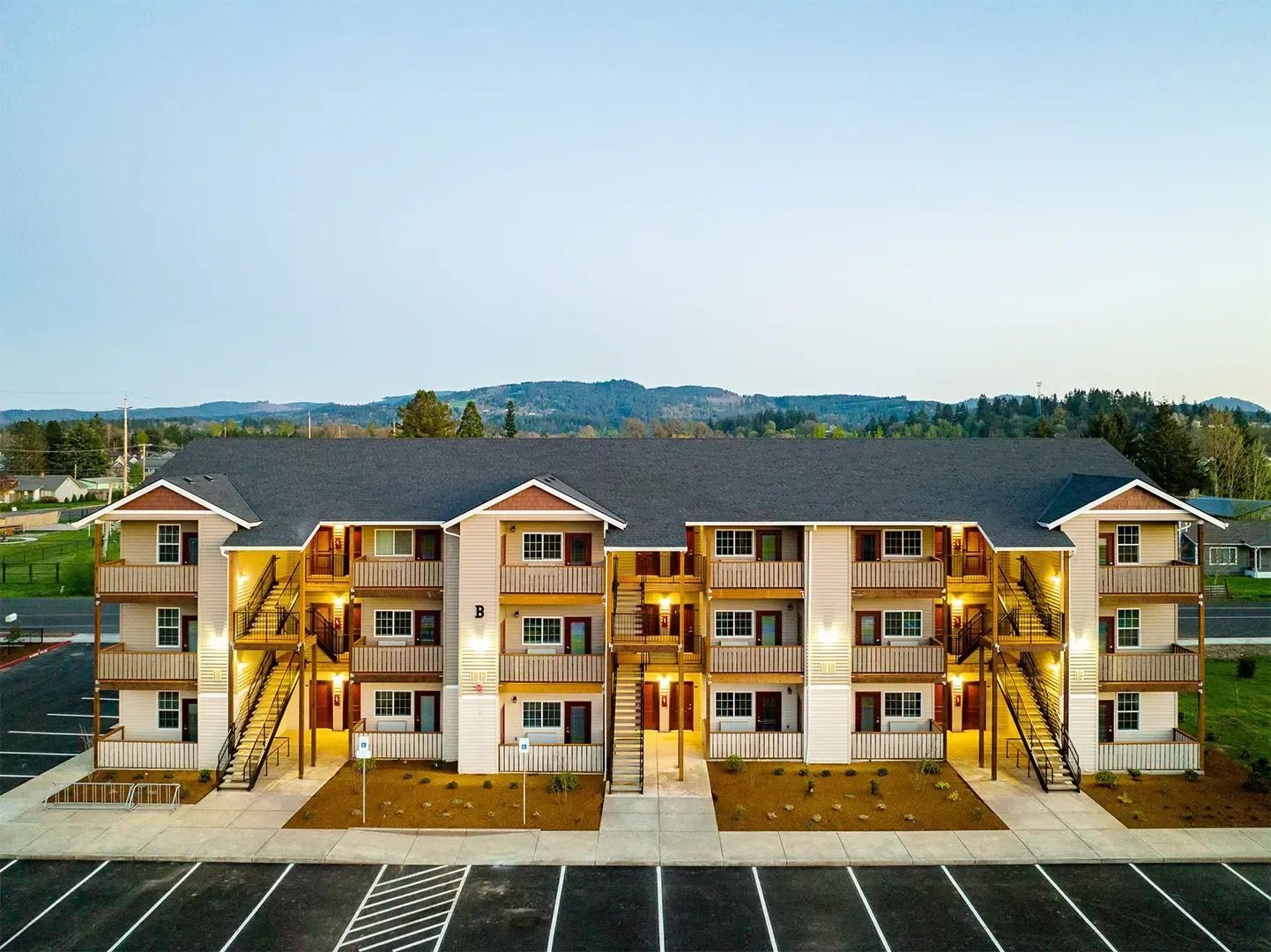 Three-story tan apartment building with exterior staircases and balconies, viewed from a parking lot.