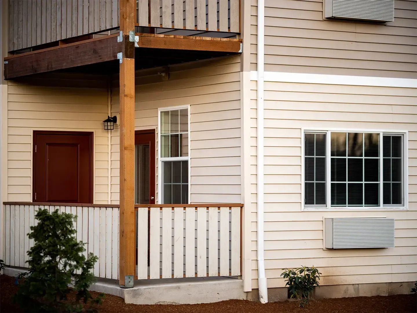 Apartment building exterior with beige siding, white balcony railings, and a red door under an upper deck