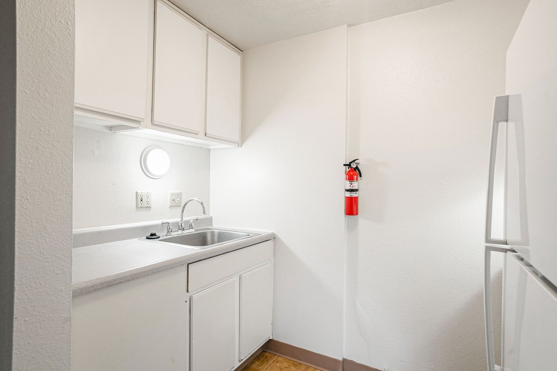 Small white kitchenette with sink, cabinets, mirror, and red fire extinguisher on the wall.