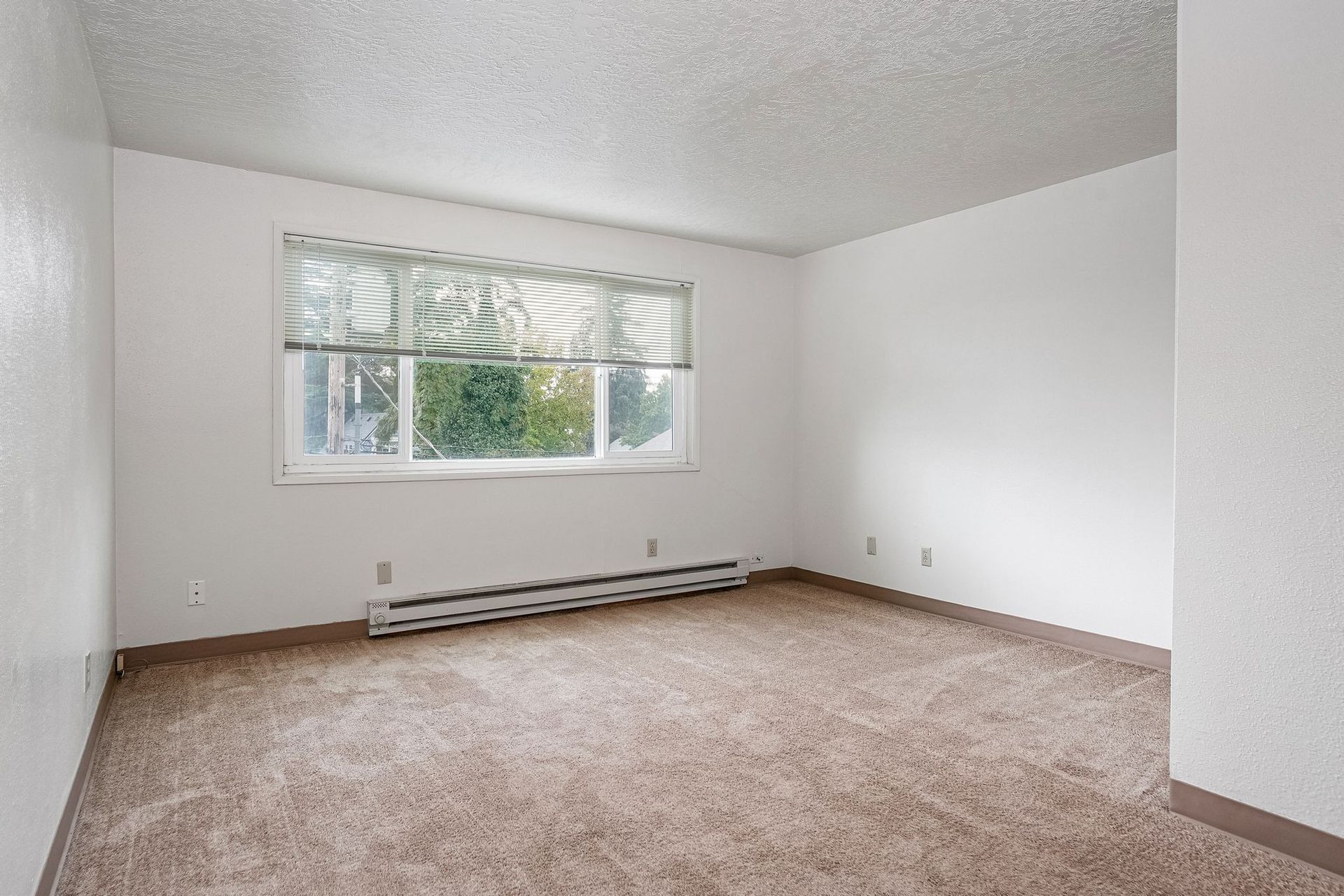 Empty carpeted living room with white walls and a window above a baseboard heater