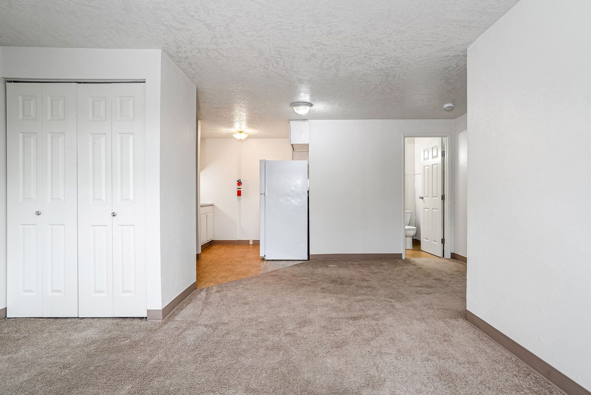 Empty carpeted apartment living room with white walls, closet doors, and kitchen visible in back
