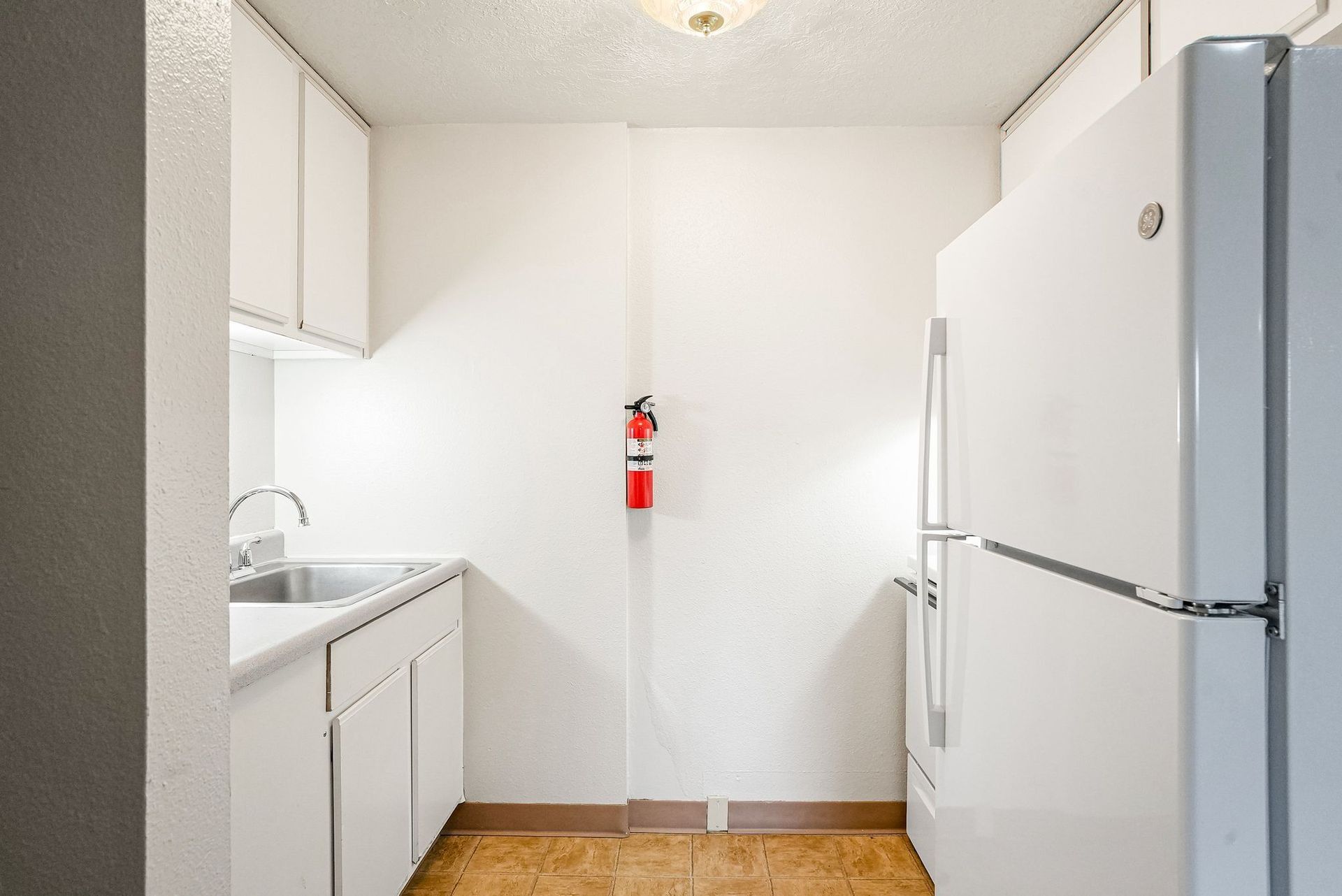 Small white kitchen with cabinets, sink, refrigerator, and a fire extinguisher on the back wall.