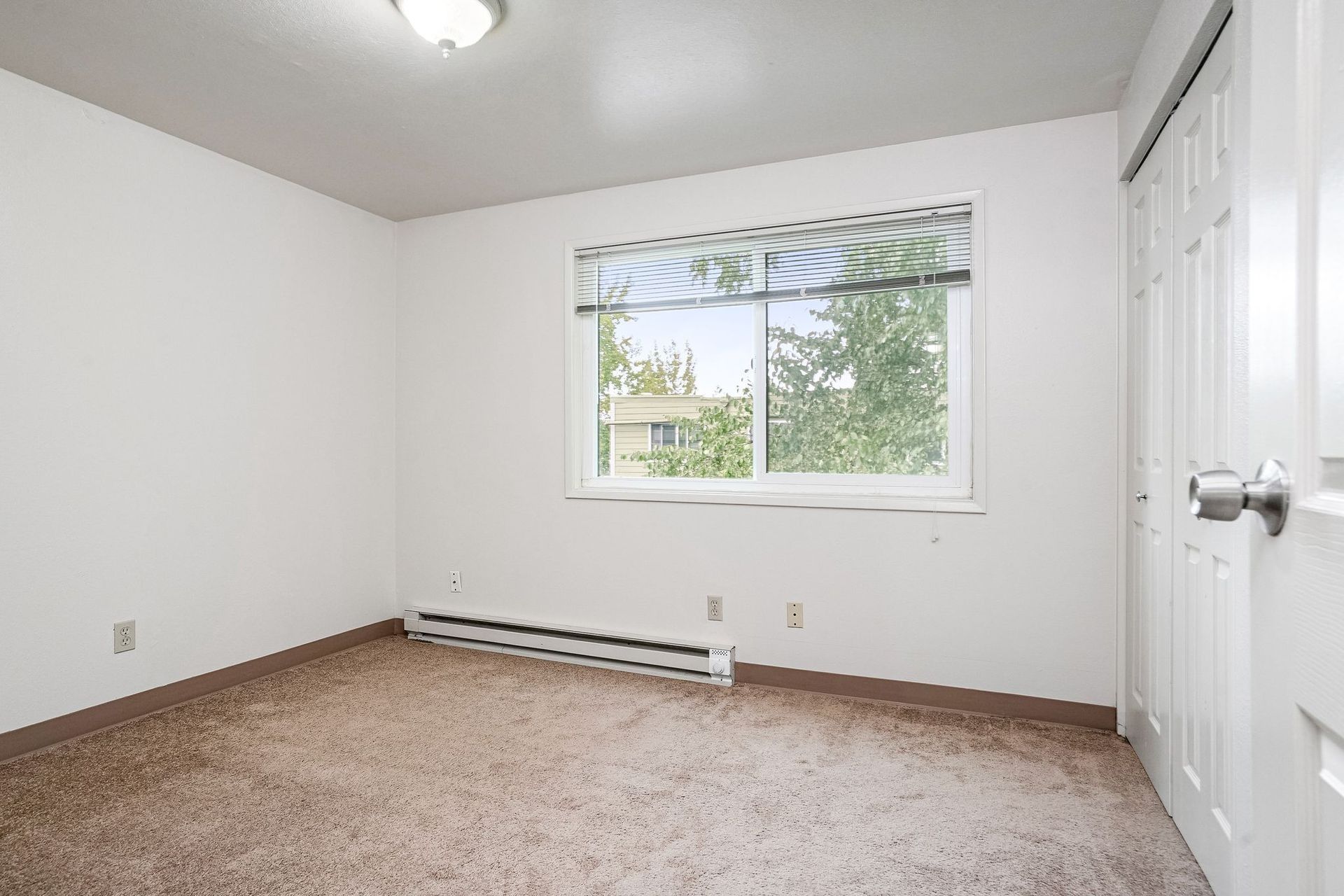 Empty beige bedroom with white walls, carpet, and a window with blinds and greenery outside