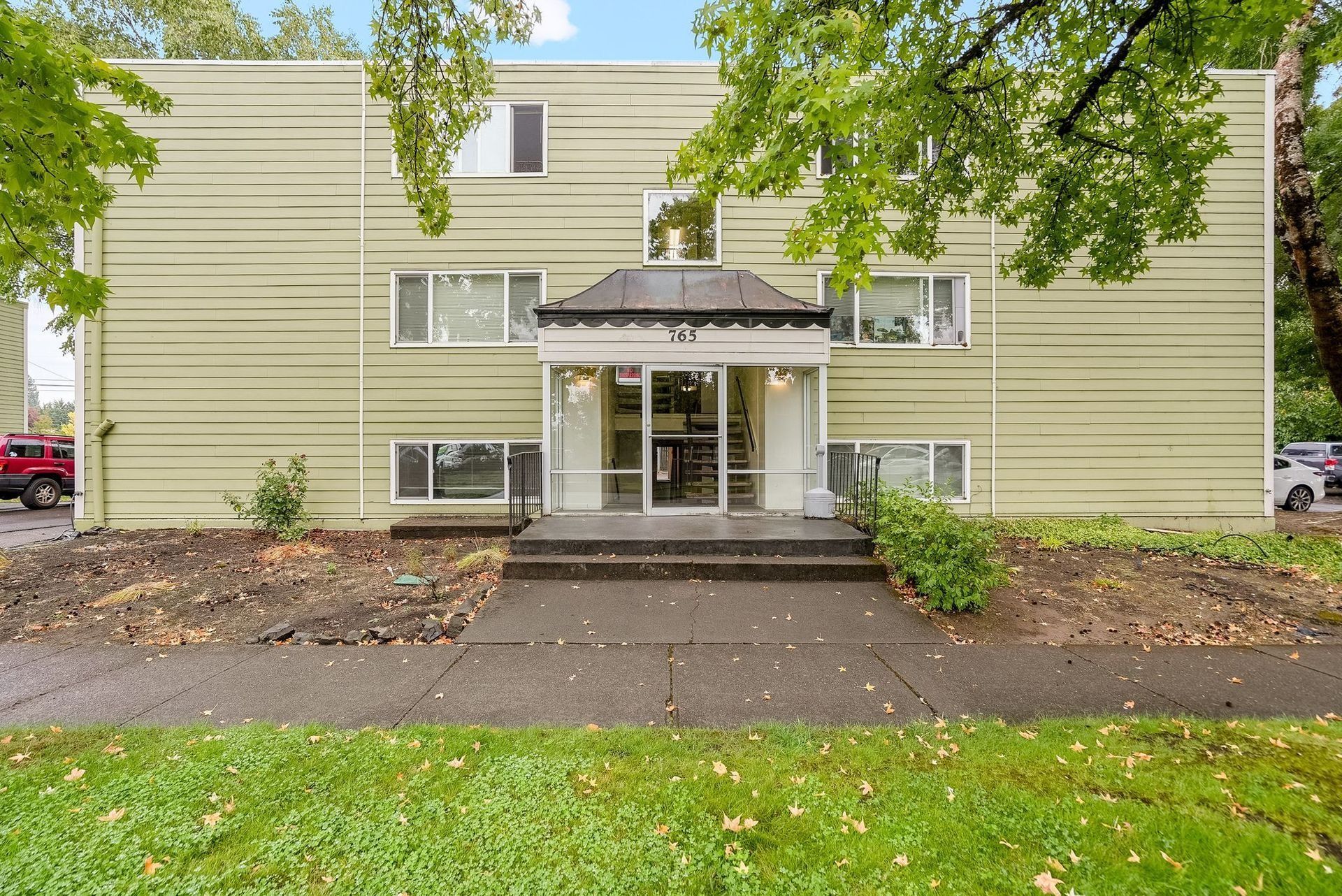 Apartment building entrance with covered steps, surrounded by trees and a grassy lawn