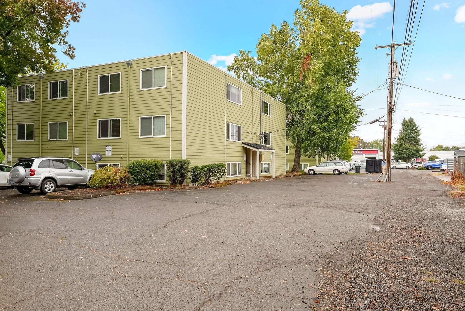 Green two-story apartment building beside a gravel lot with parked cars under a clear sky