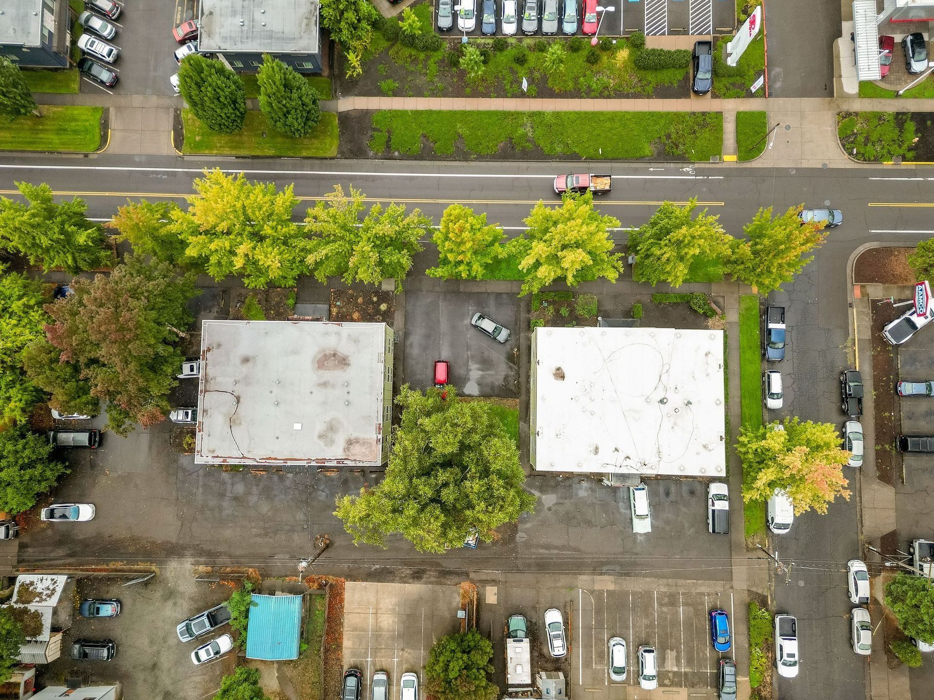 Aerial view of two white-roofed buildings beside roads and parked cars, surrounded by trees and parking lots