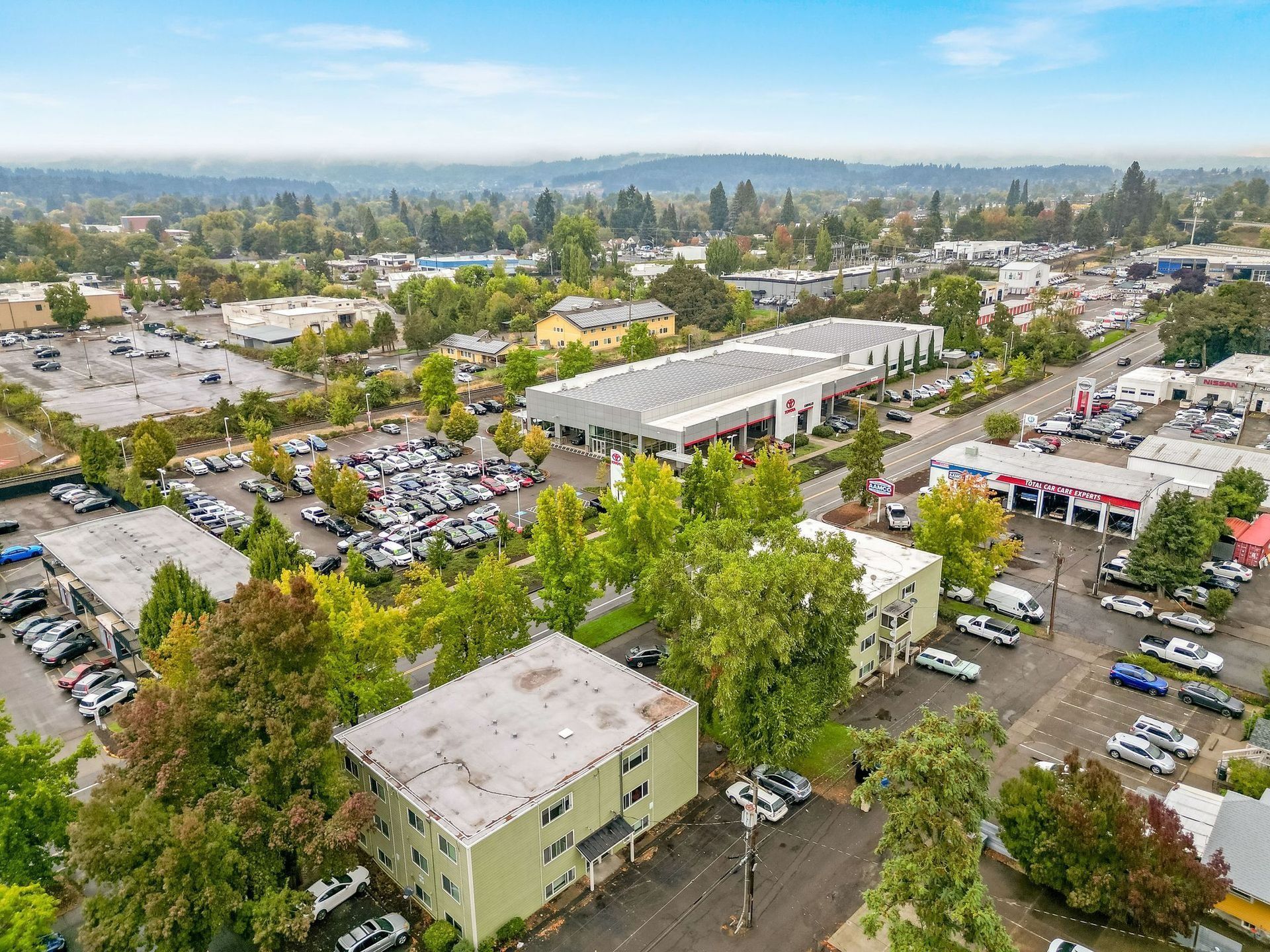 Aerial view of a suburban shopping center with parking lots, trees, and surrounding low-rise buildings.