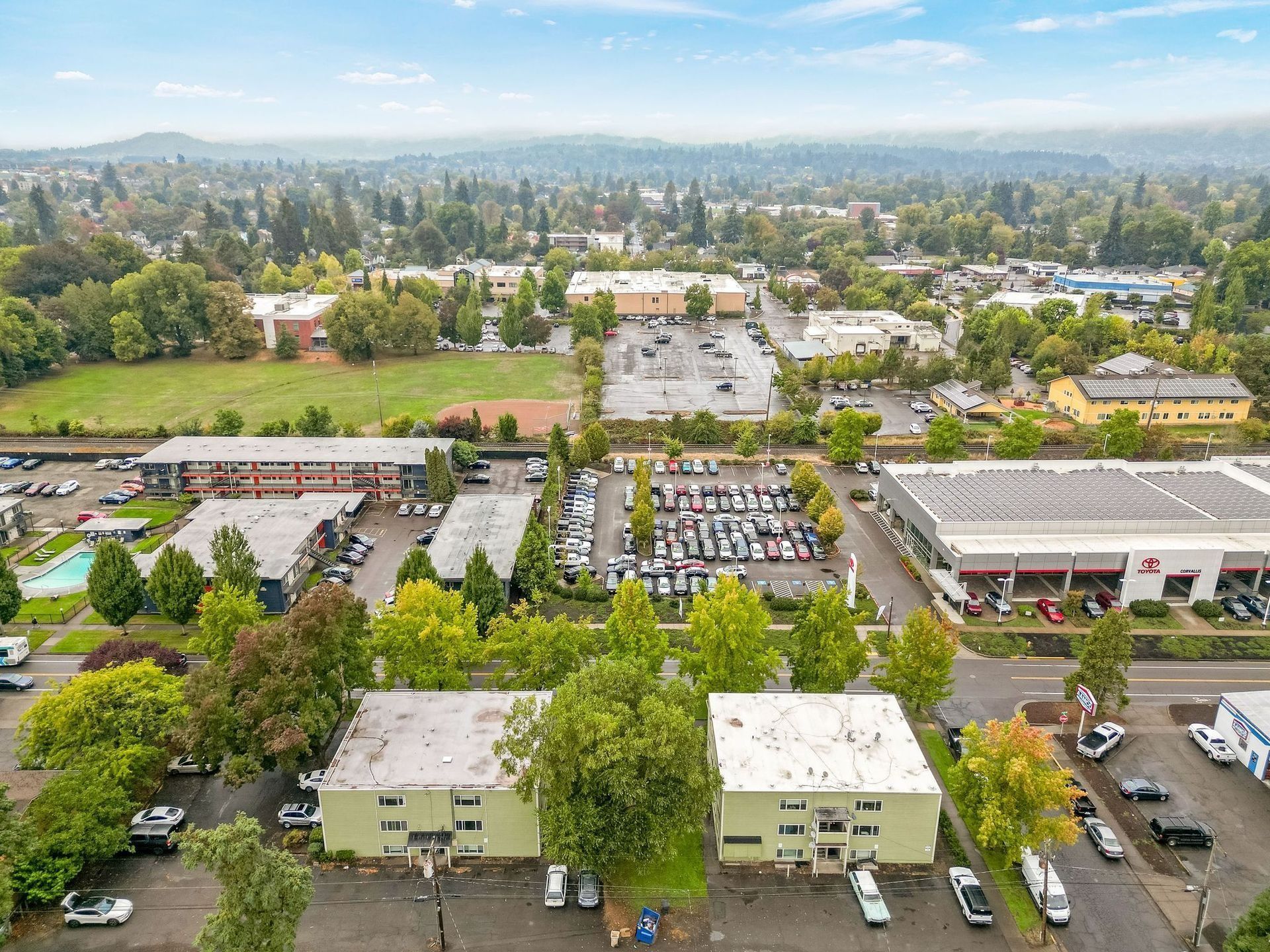 Aerial view of a suburban shopping center with parking lots, trees, and low-rise buildings under a hazy sky