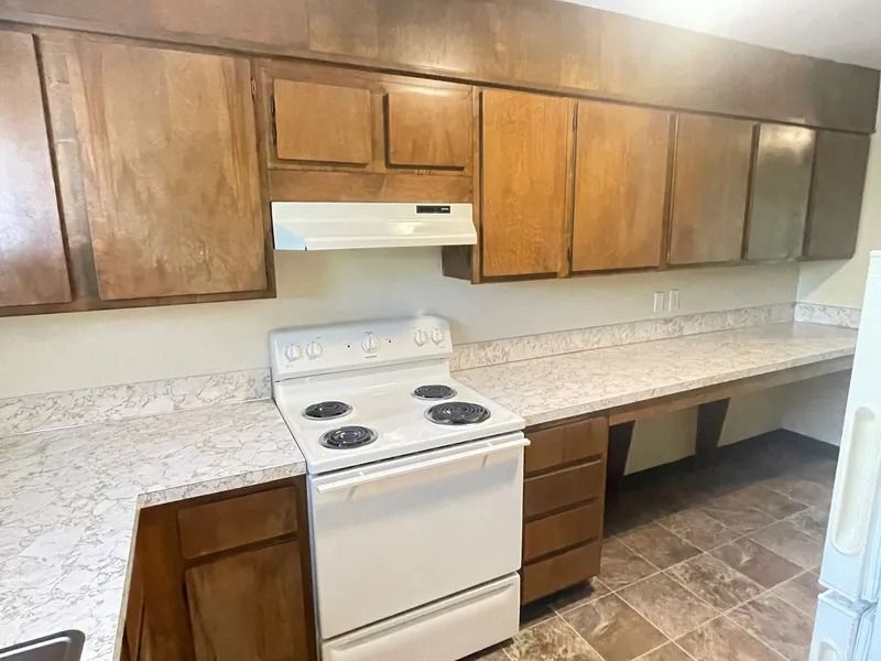 Empty kitchen with wooden cabinets, white stove, and long speckled countertop under overhead lighting