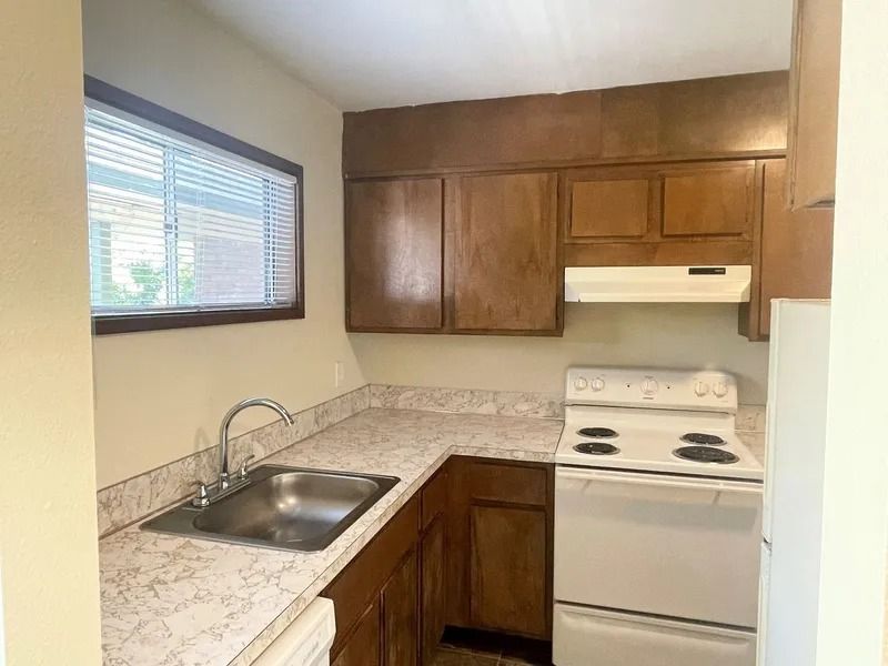 Small kitchen with wood cabinets, white stove, sink under a window, and beige countertops.