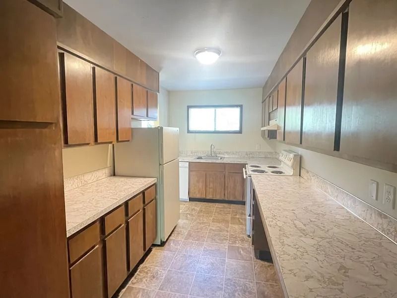 Narrow kitchen with wooden cabinets, white appliances, beige counters, and a window at the far end.