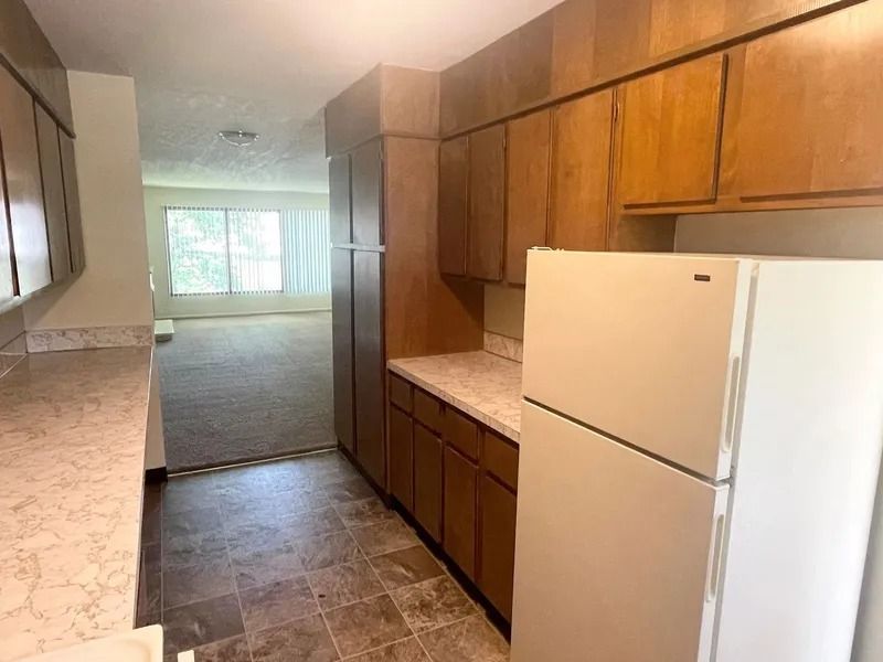 Narrow kitchen with wood cabinets, white refrigerator, and a view into a carpeted living room.