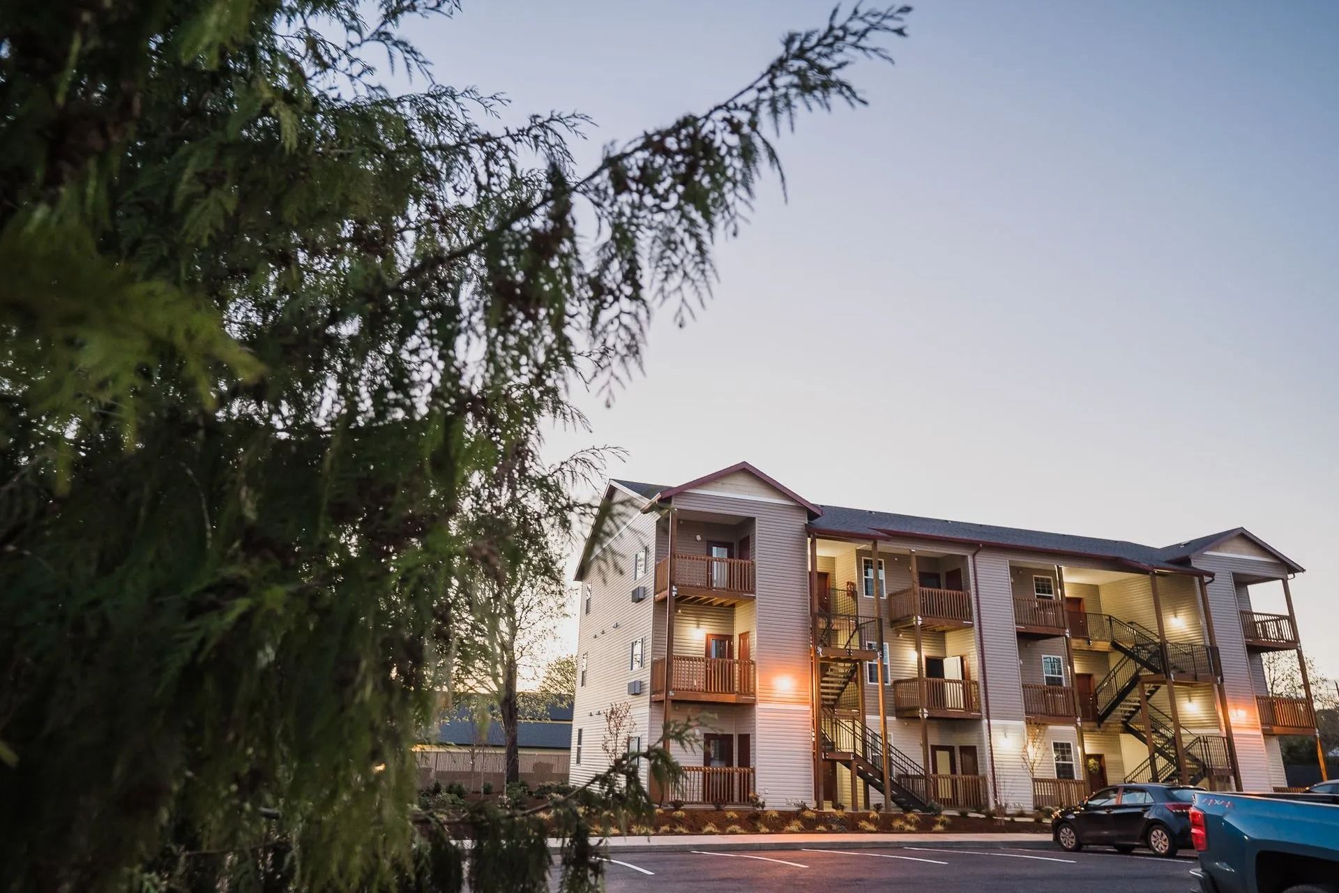 Apartment building at dusk, framed by tree branches, with lit balconies and parked cars in front.