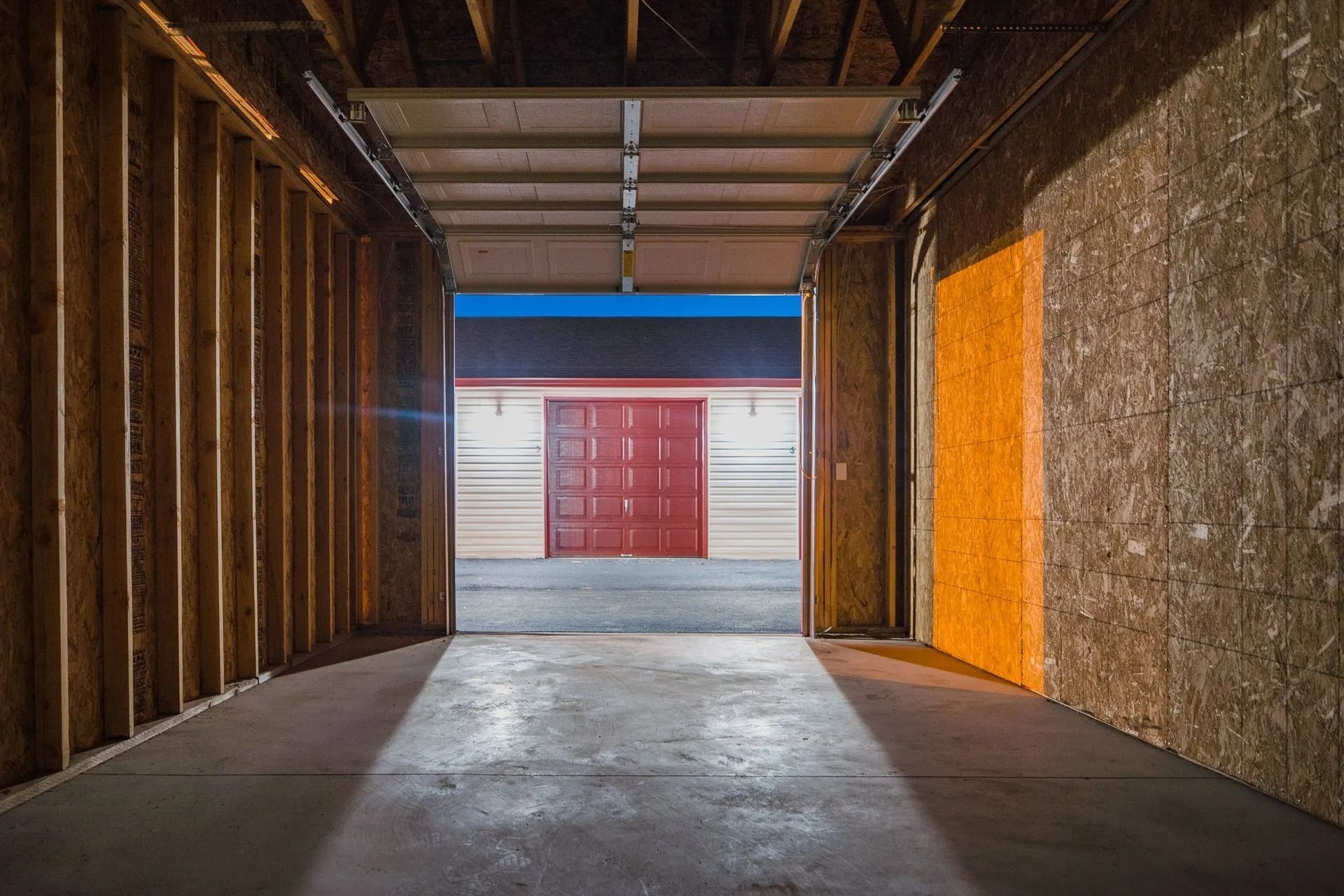Empty warehouse interior with a concrete floor, wood-framed walls, and an open doorway to a bright red building.