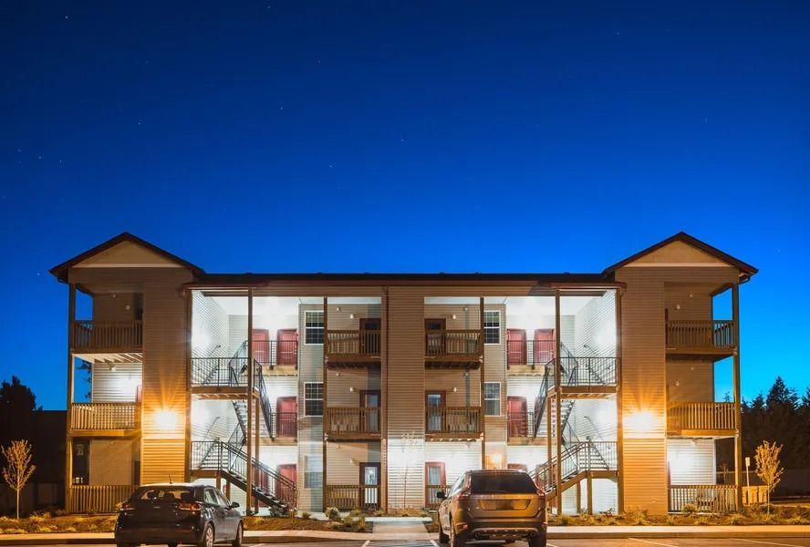 Night view of a three-story apartment building with lit windows and parked cars in front
