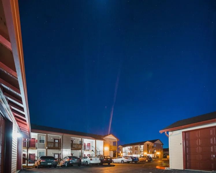 Nighttime apartment complex parking lot with lit buildings, cars, and a blue sky with a streak of light.