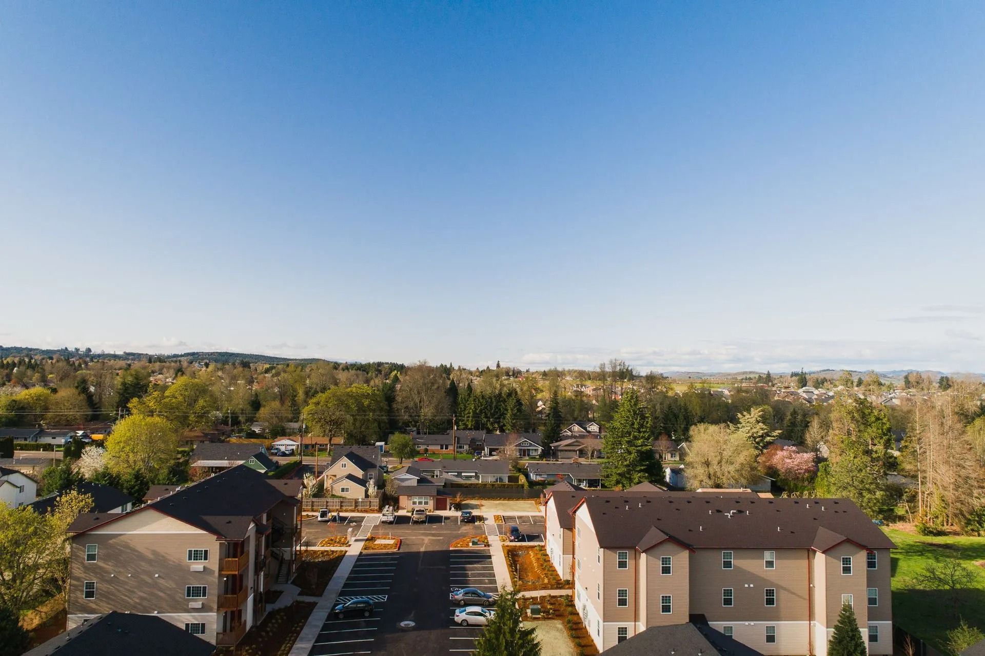 Aerial view of apartment buildings and a parking lot under a clear blue sky with trees in the background