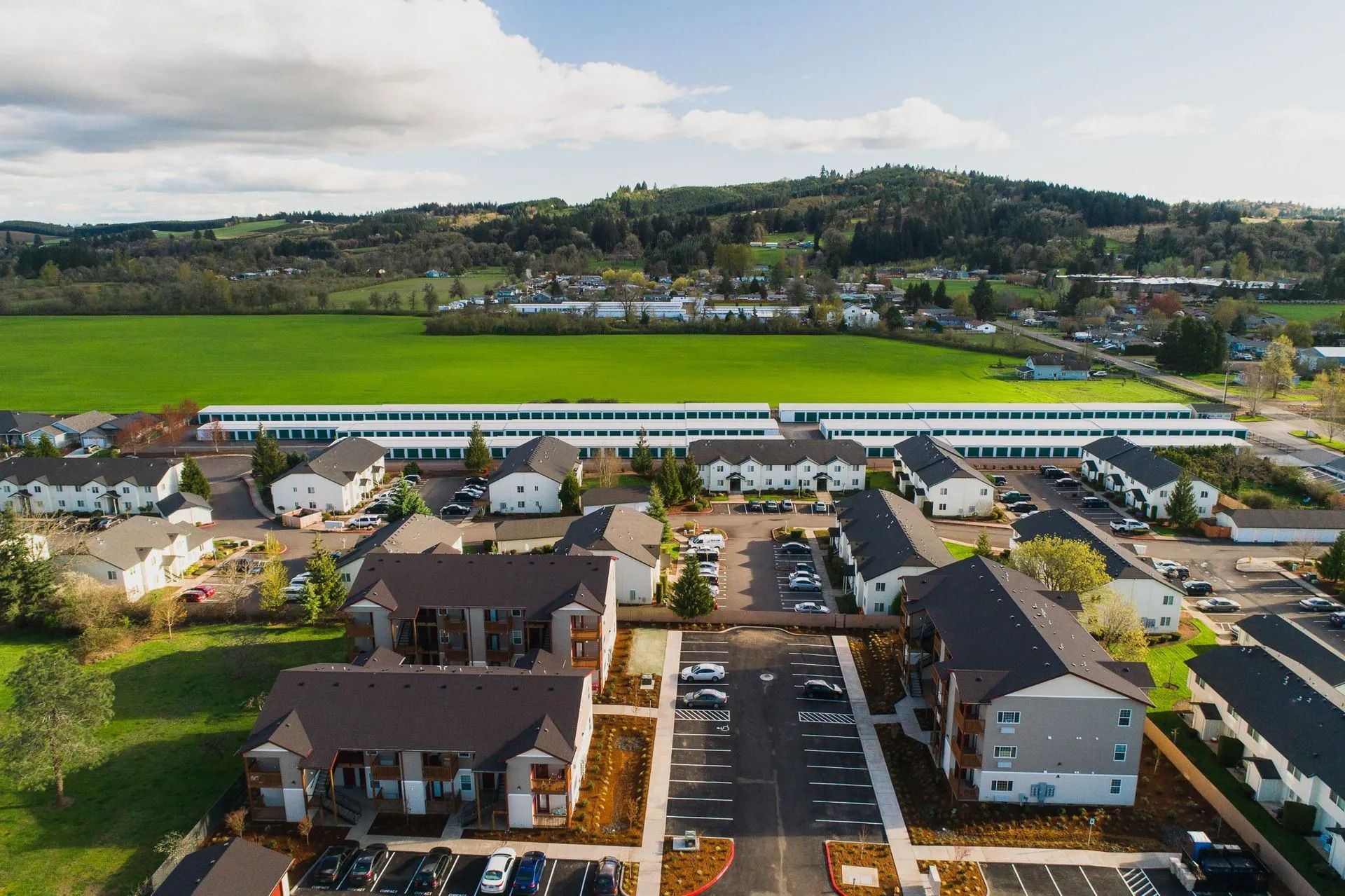 Aerial view of a suburban neighborhood with homes, parking lots, fields, and wooded hills in the background