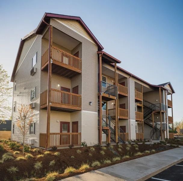 Three-story apartment building with wooden balconies and beige siding along a sidewalk