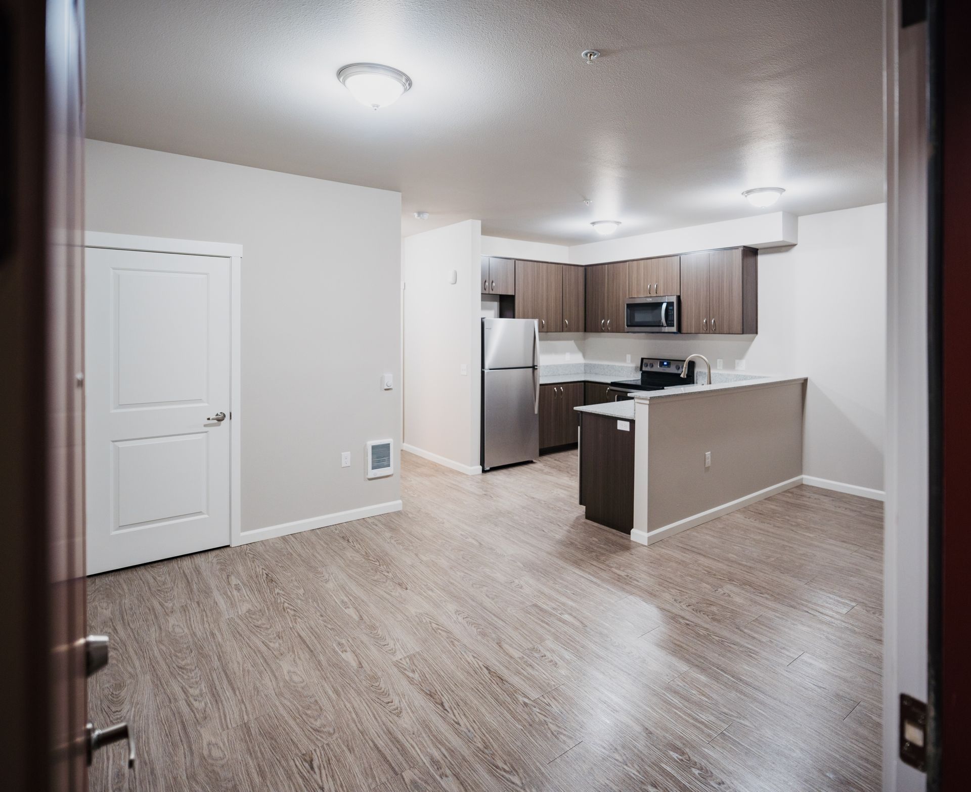 Empty modern apartment interior with white walls, wood-look flooring, and a compact kitchen with gray cabinets and island