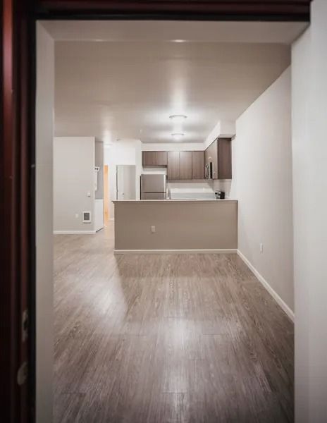 Open-concept apartment with light wood floors, white walls, and a kitchen visible beyond a half wall.