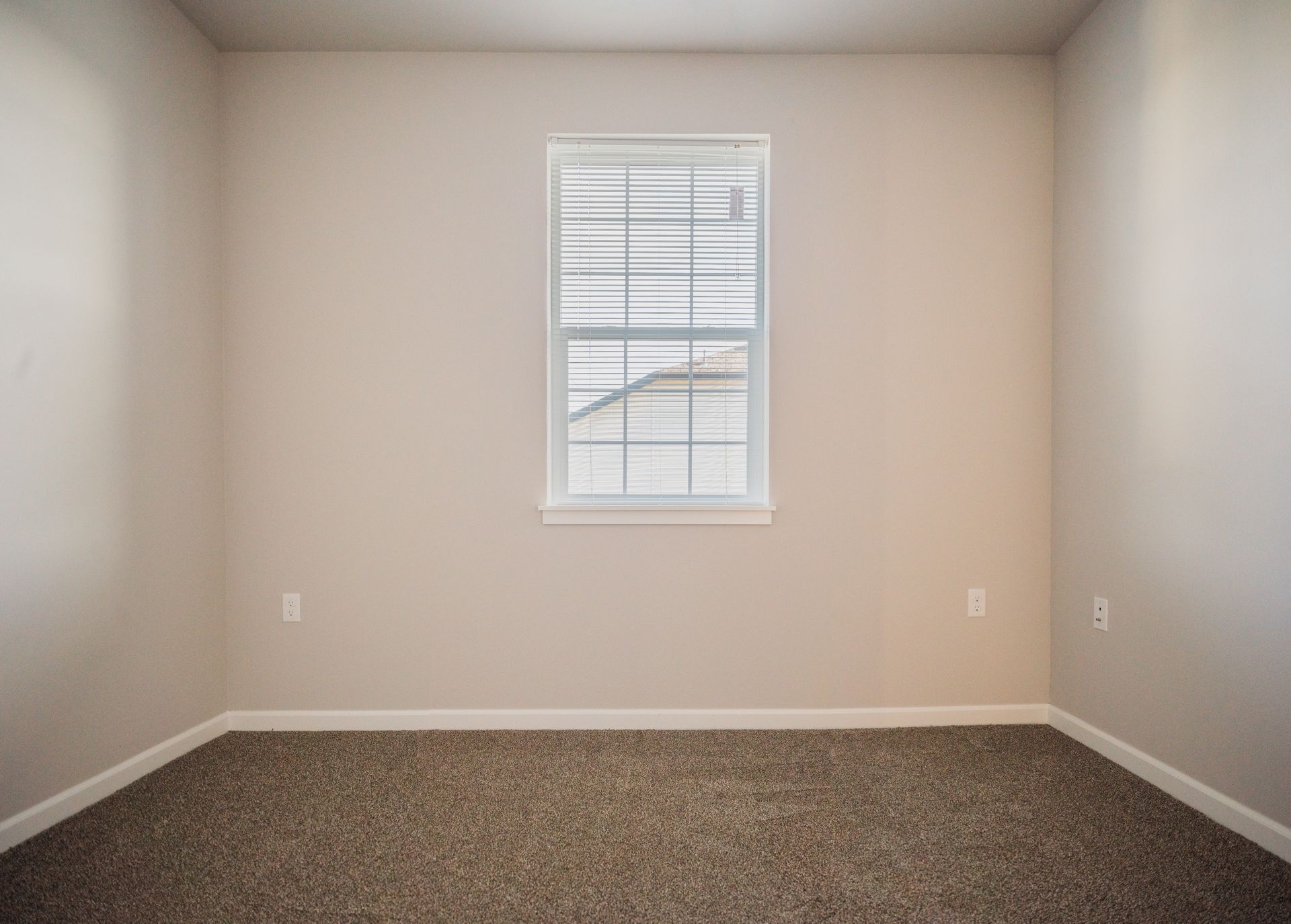 Empty carpeted room with beige walls and a small window centered on the far wall
